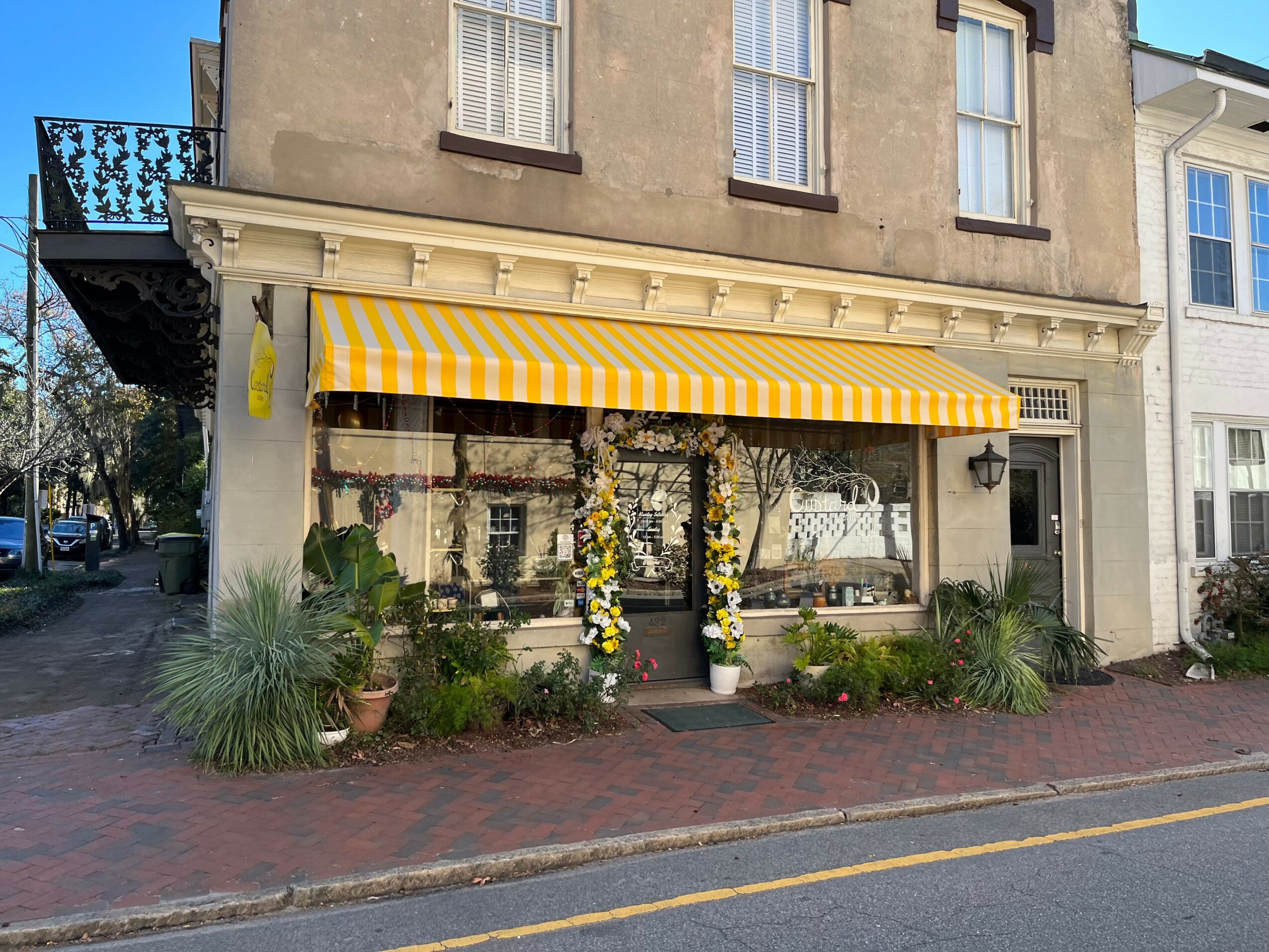 Storefront with yellow and white striped awning, plants, and flowers outside, in a building on a city street.