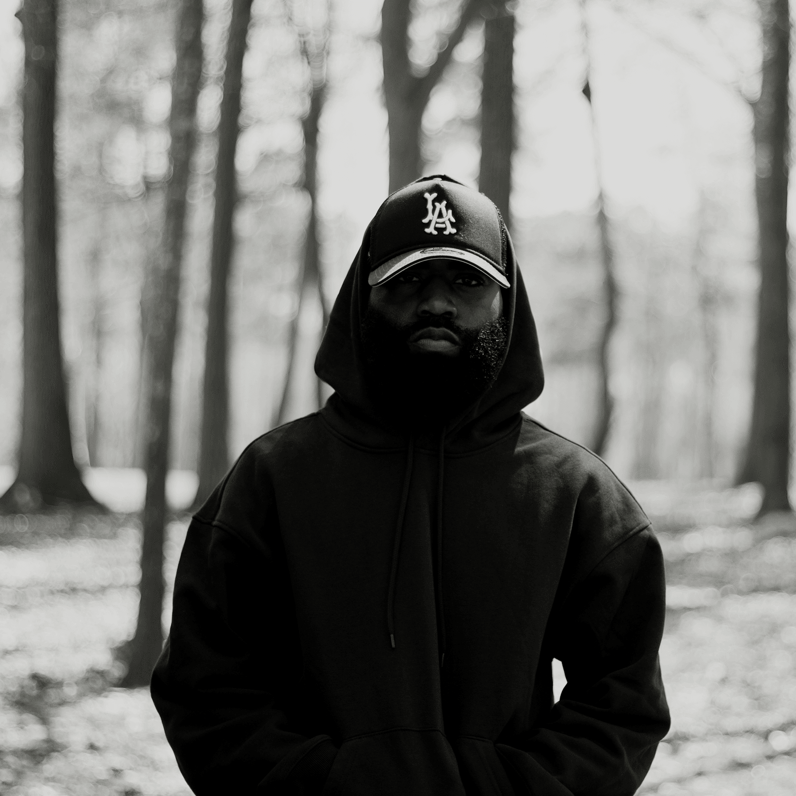 Person wearing a hoodie and cap standing in a forest with trees in the background.