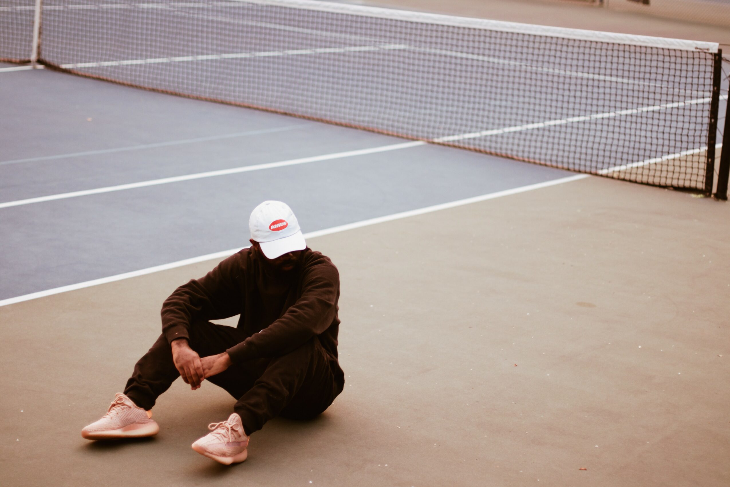 Person sitting on the ground near a tennis court, wearing a white cap, dark clothing, and pink shoes.