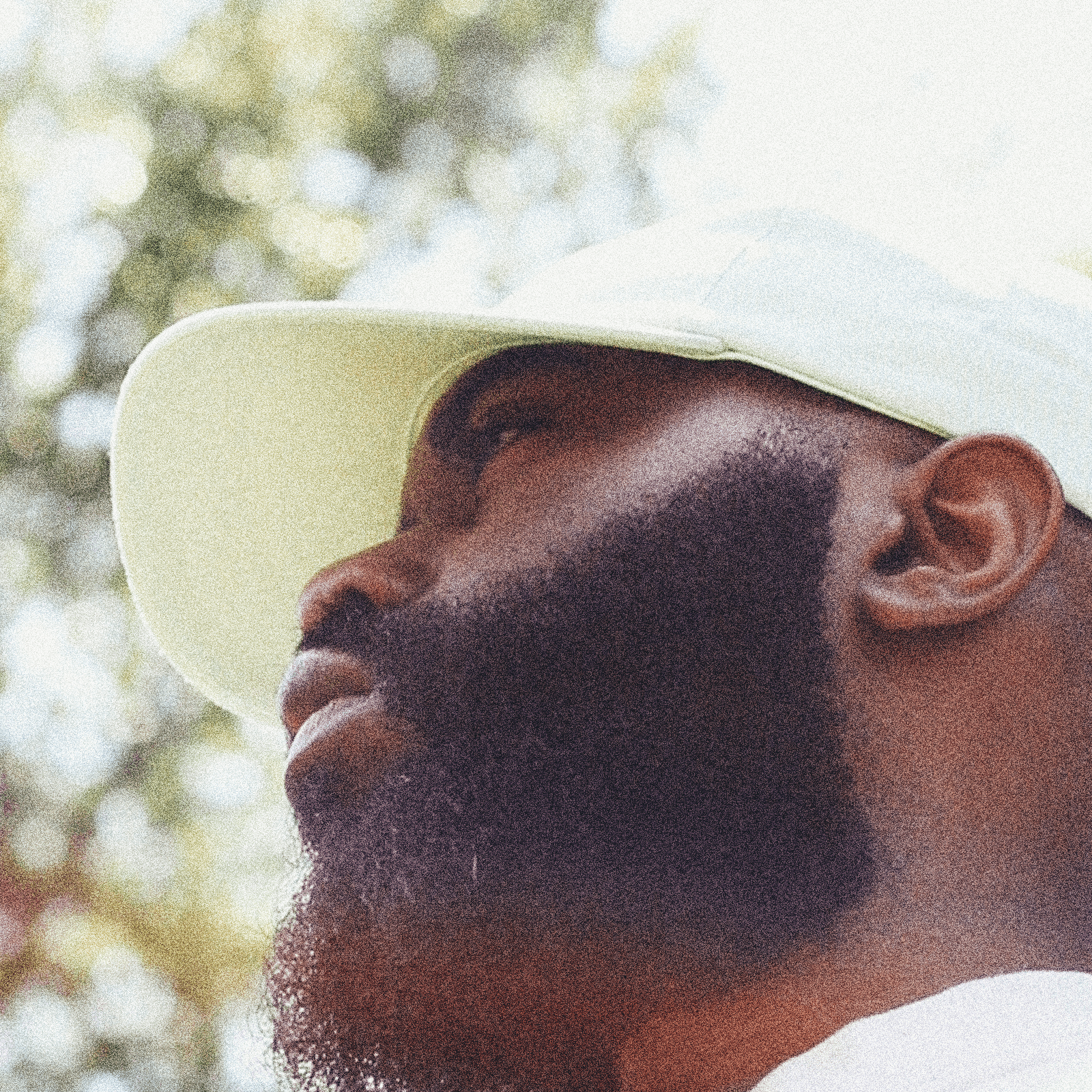 Close-up of a man with a beard wearing a white hat outdoors, with trees in the background.