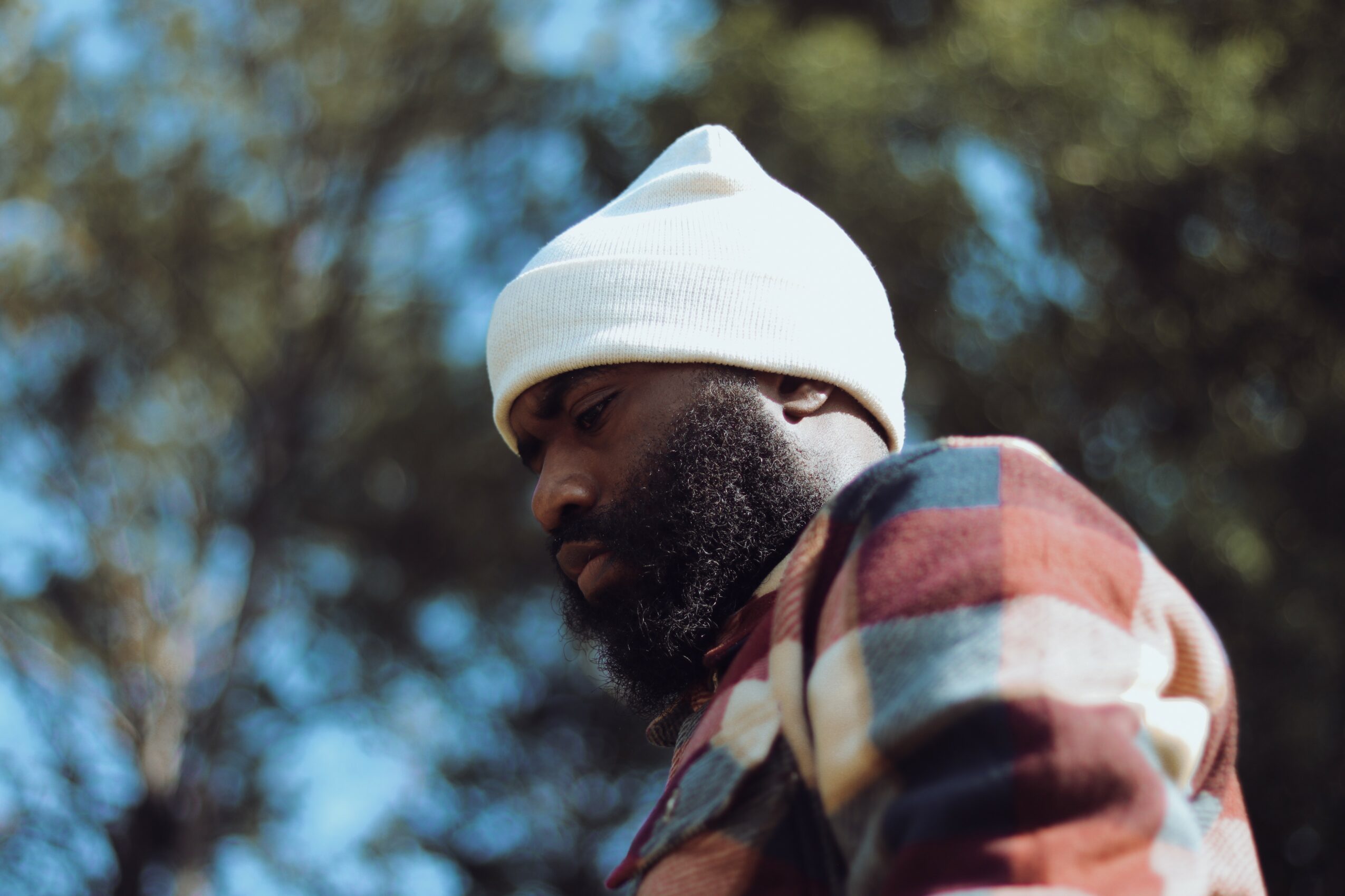 Man with a beard wearing a white beanie and a multicolored jacket outdoors, trees in background.