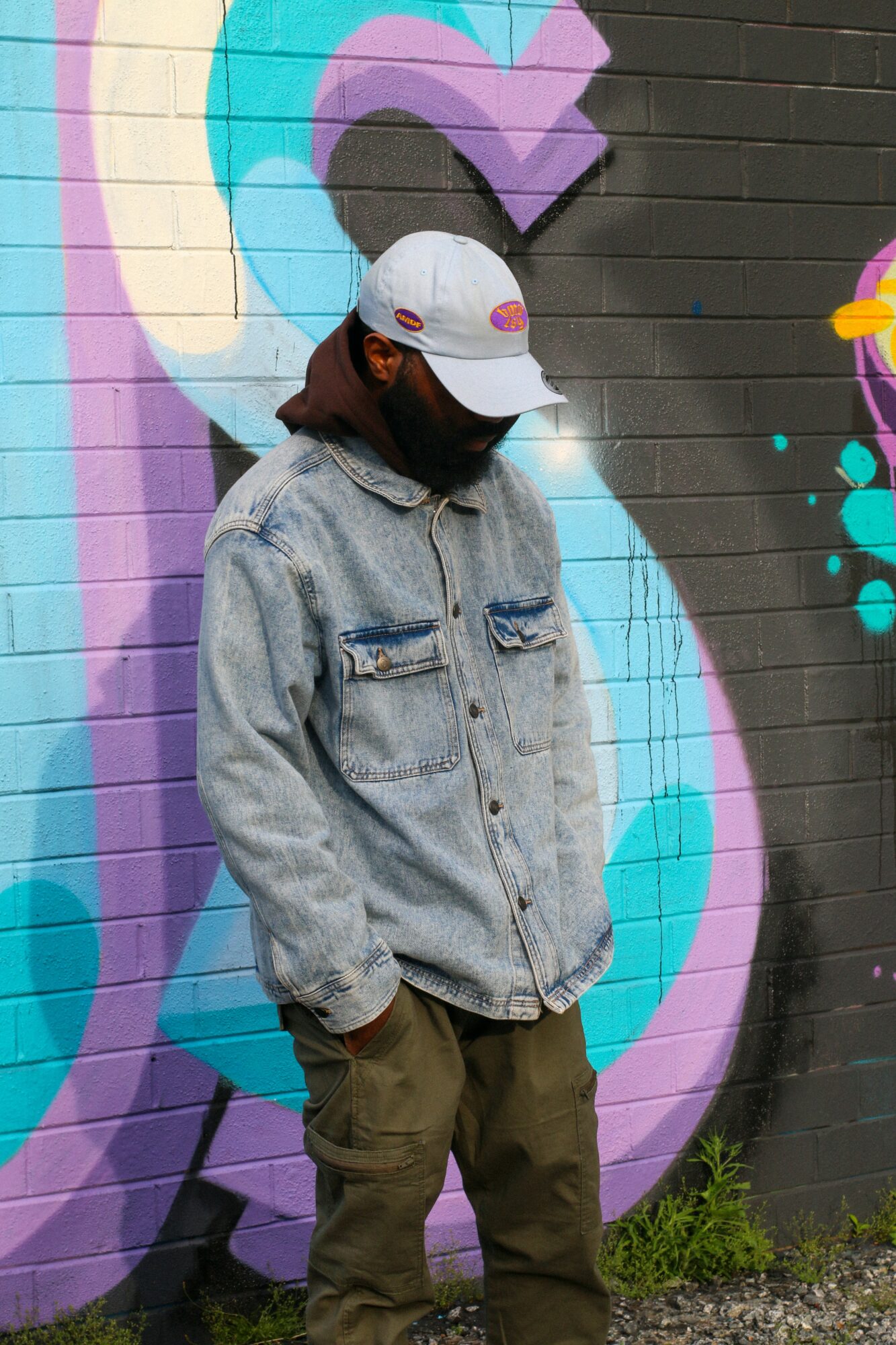 Person wearing a white cap and denim jacket stands against a colorful graffiti wall.