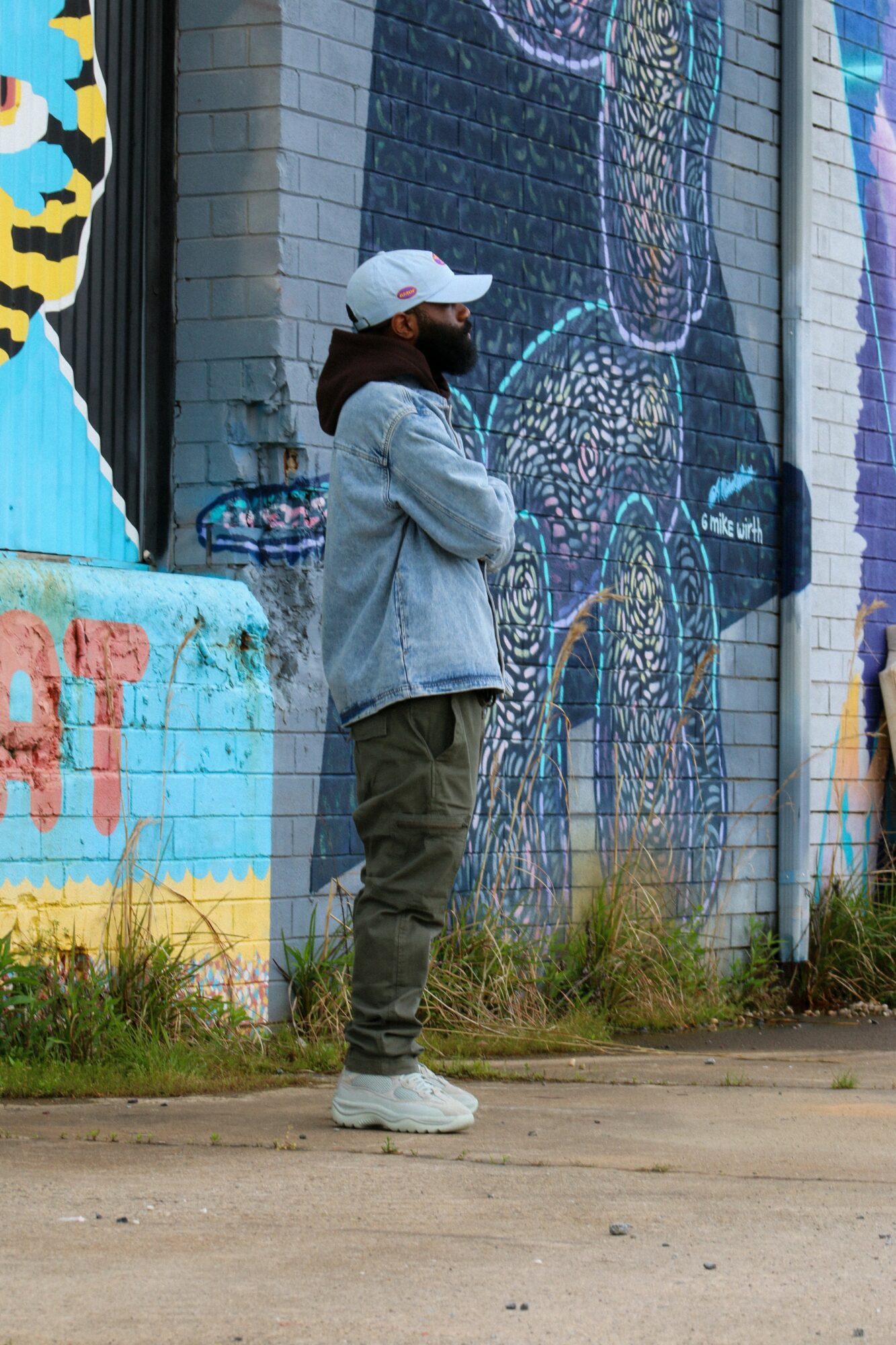 Person standing in front of colorful mural, wearing a white cap, denim jacket, and cargo pants, with grass and sidewalk nearby.