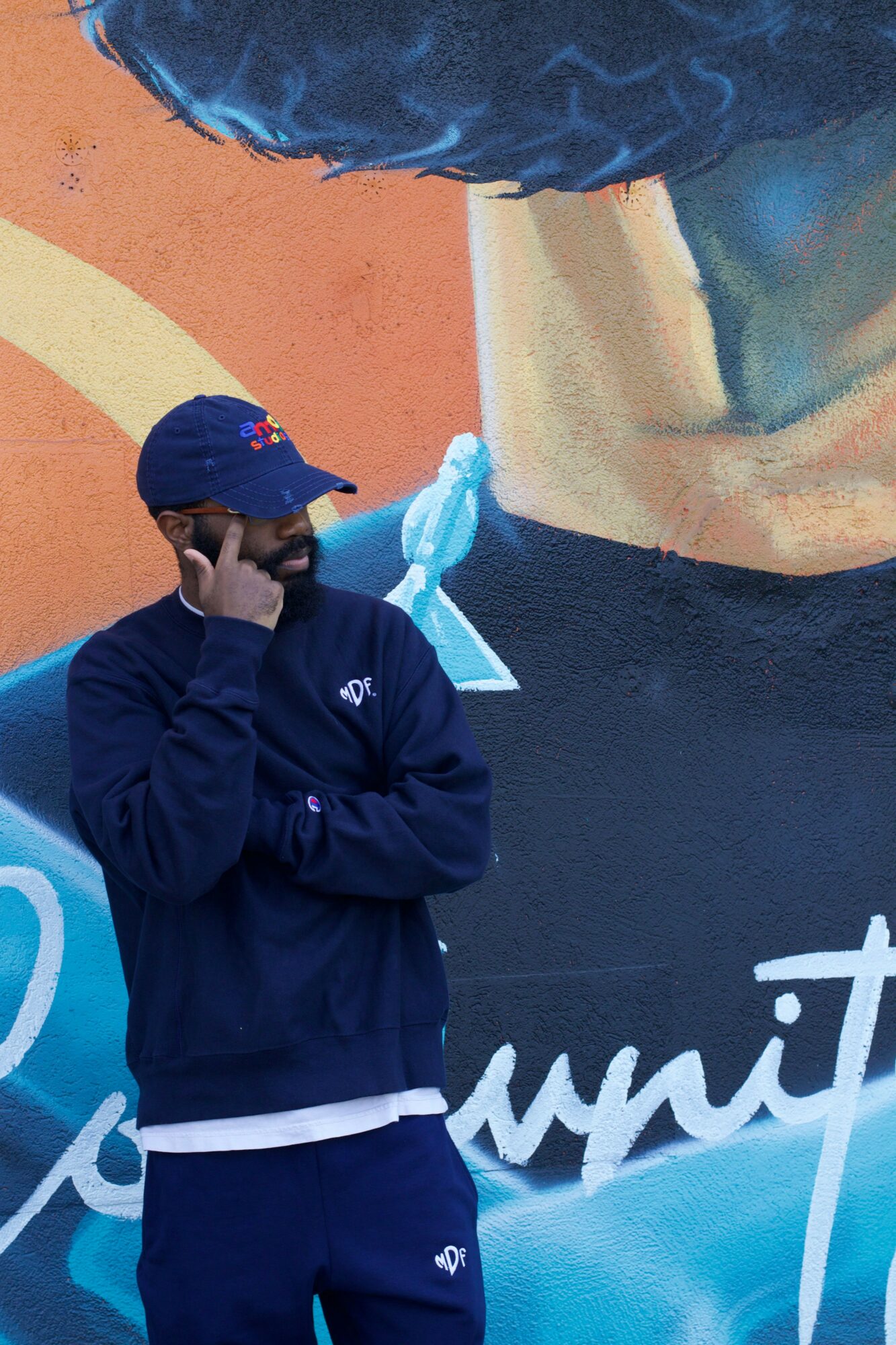 Person wearing a dark blue jacket and a blue cap, standing outdoors near a wall with orange and beige colors.