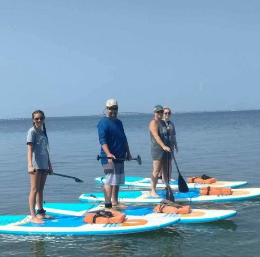 Four people stand on paddleboards in the water, facing the camera, with a clear sky and ocean background.