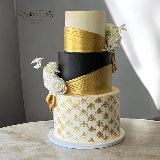 Three-tiered cake with gold, black, and white decorations, adorned with white flowers and gold patterns, on a table.