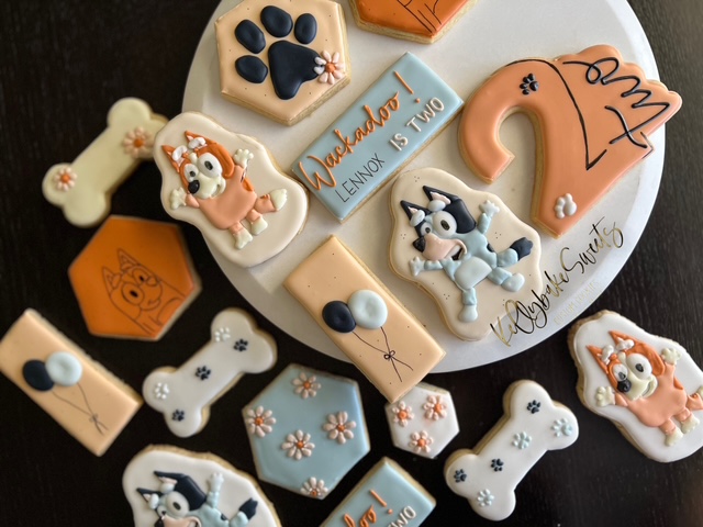 Decorated cookies with dog and bone shapes, a birthday cake, and a sign for a child's birthday party, on a white plate.