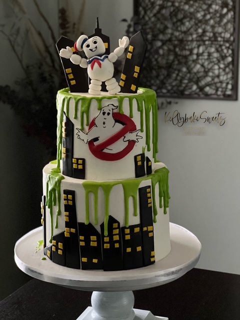 Two-tier cake with Ghostbusters theme, featuring a cartoon ghost and a character on top, green drips, and cityscape decorations, on a white cake stand.