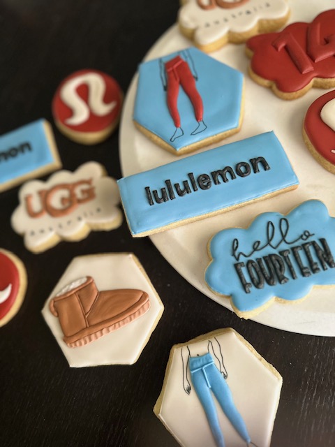 Assorted decorated cookies with clothing and brand logos, including jeans, UGG, Lululemon, and Hello Fourteen, on a black surface.