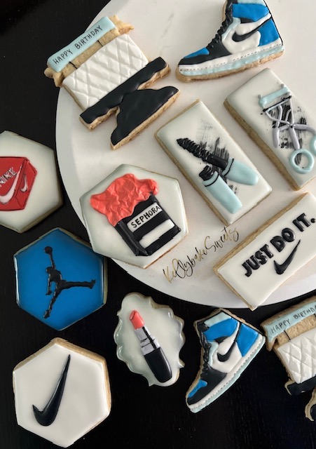 Decorative cookies shaped like sneakers, a lipstick, and a Nike swoosh, arranged on a white plate with a black background.