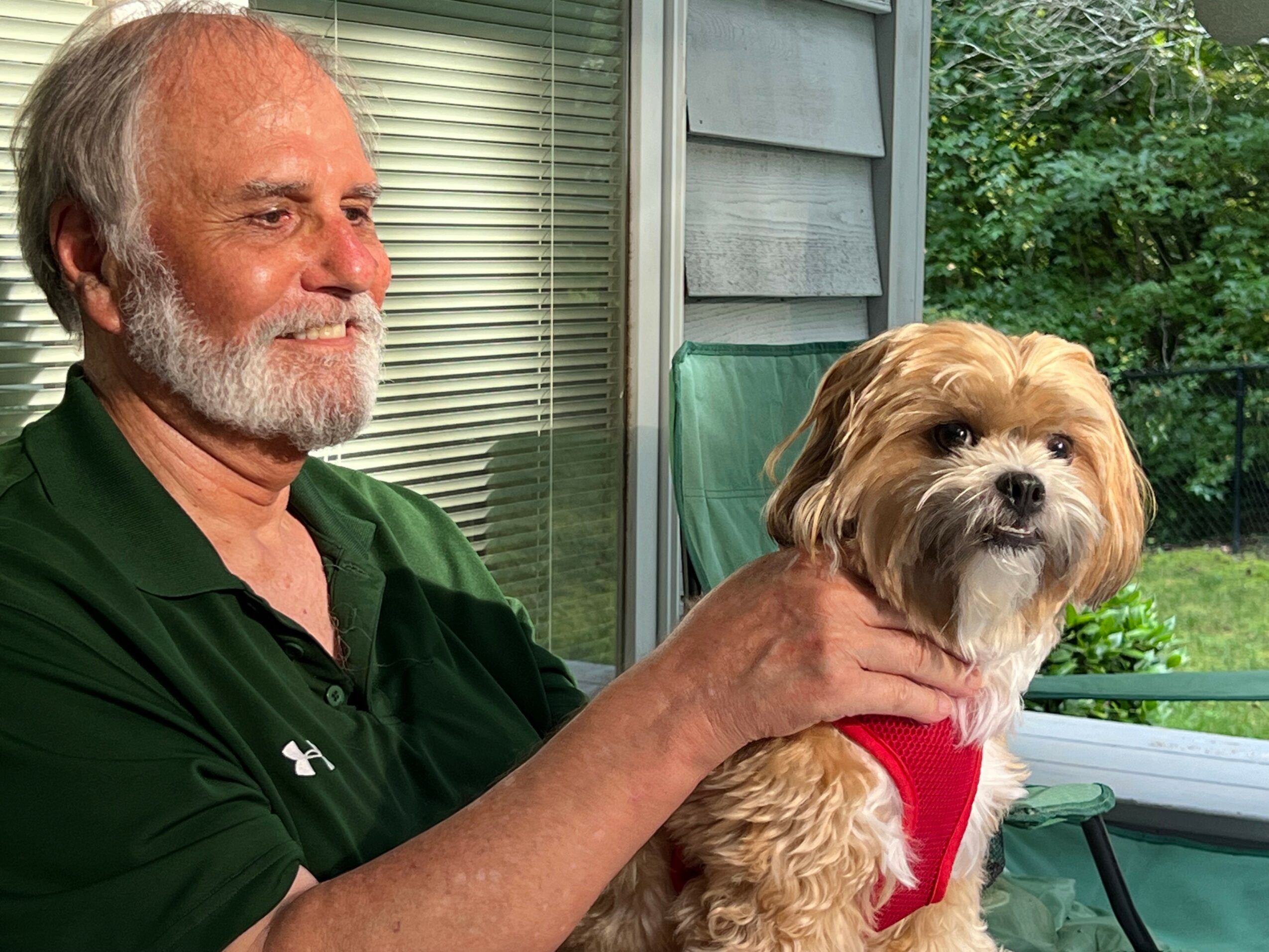Older man with gray hair and beard sitting outside with a small, fluffy dog wearing a red harness, on a porch with greenery.