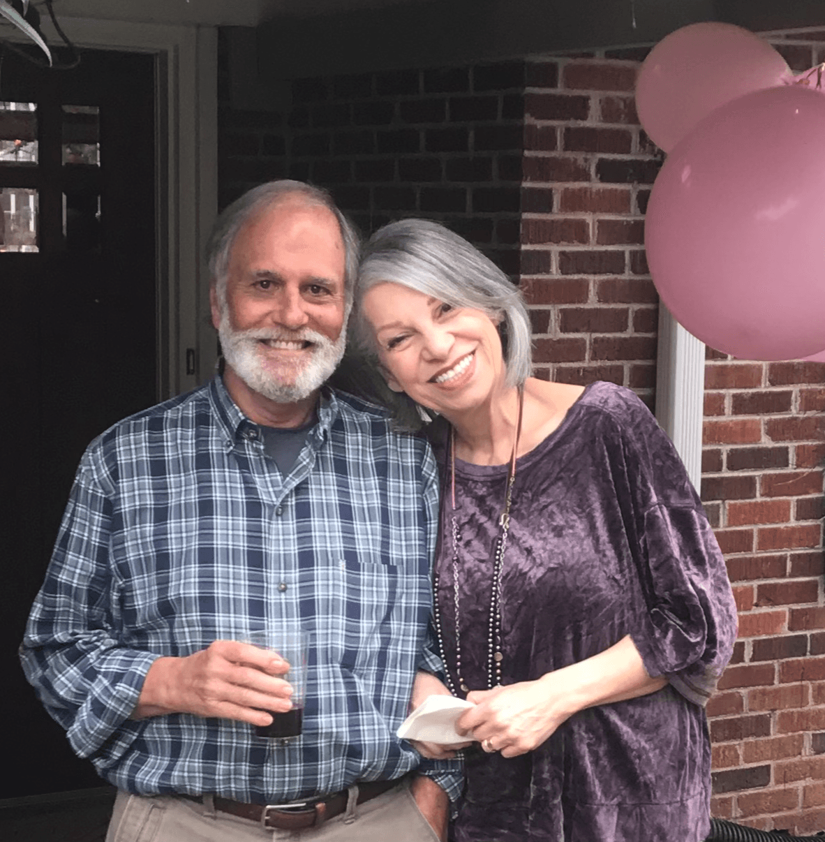 Two smiling people, a man with a beard and a woman with gray hair, stand close together in front of a brick wall, holding a small object. Pink balloons are visible in the background.