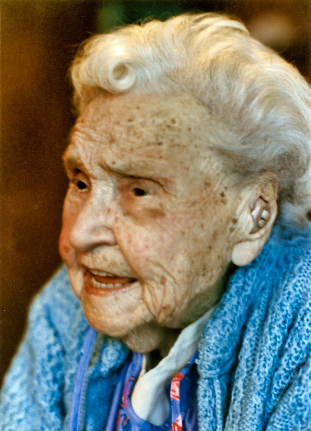 Elderly woman with white hair smiling, wearing a blue sweater, in a close-up profile view.