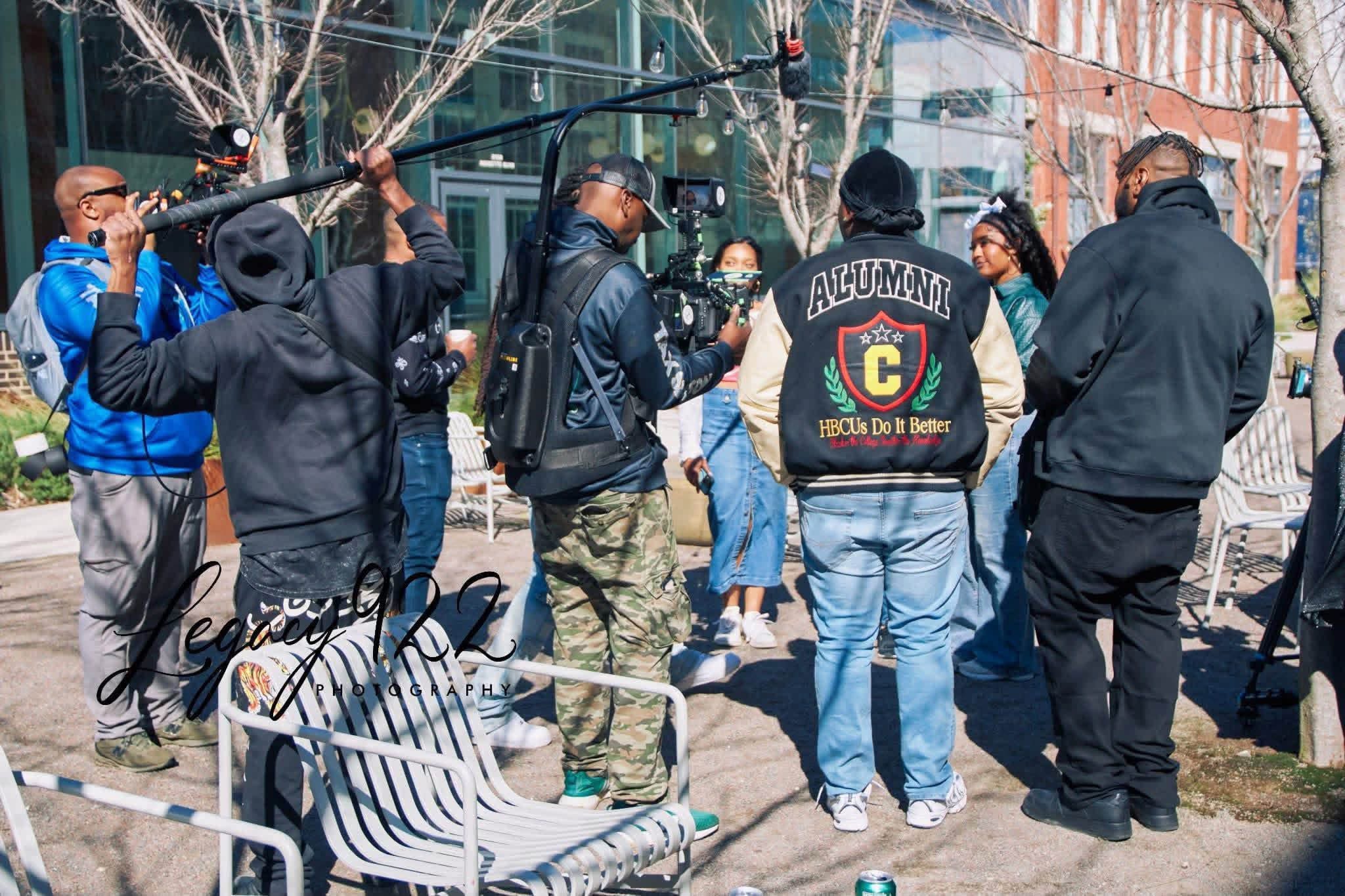 Group of people gathered outdoors, some holding microphones and equipment, with trees and buildings in background.