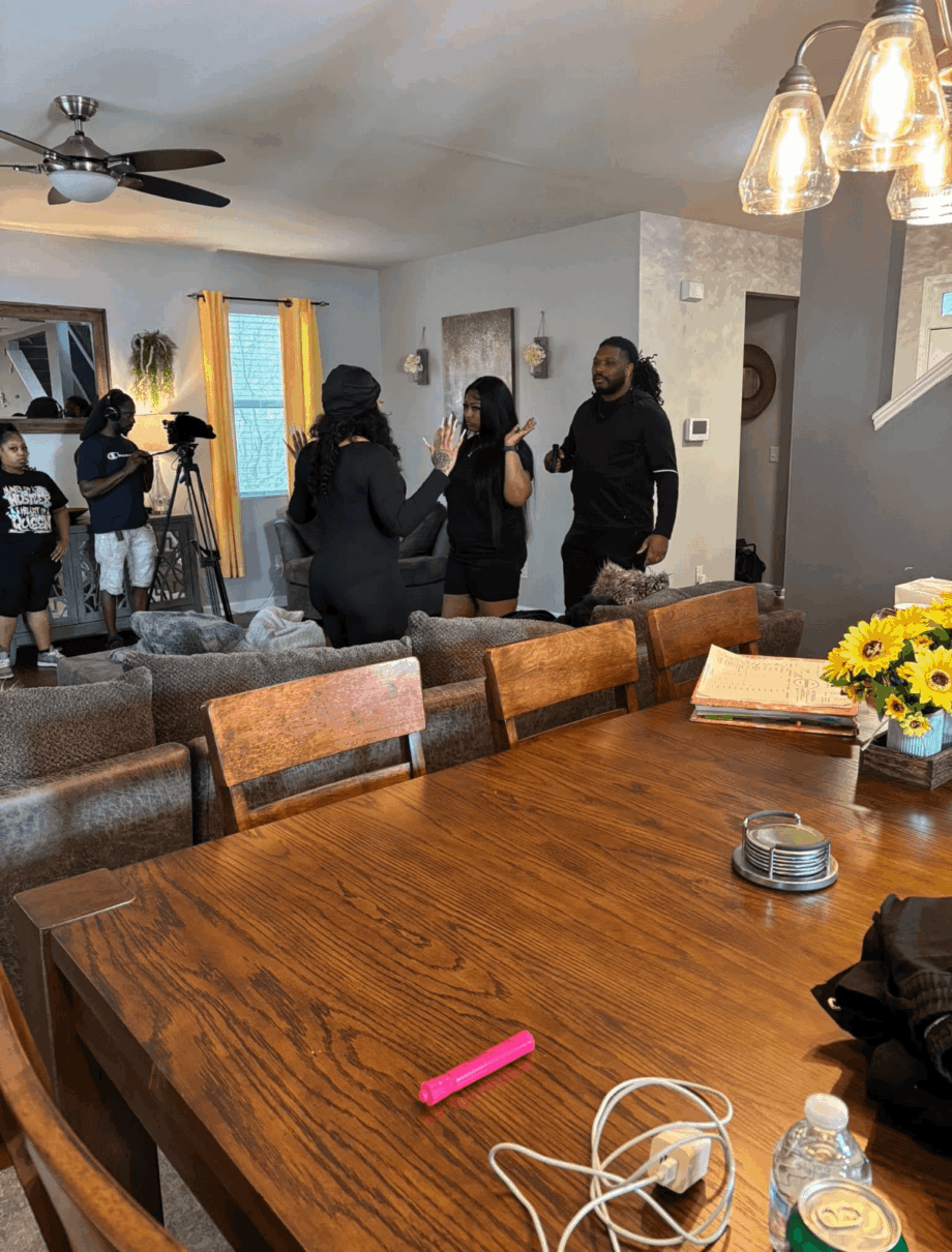 People gather in a living room with a wooden dining table, hanging lights, and a ceiling fan, engaging in conversation.