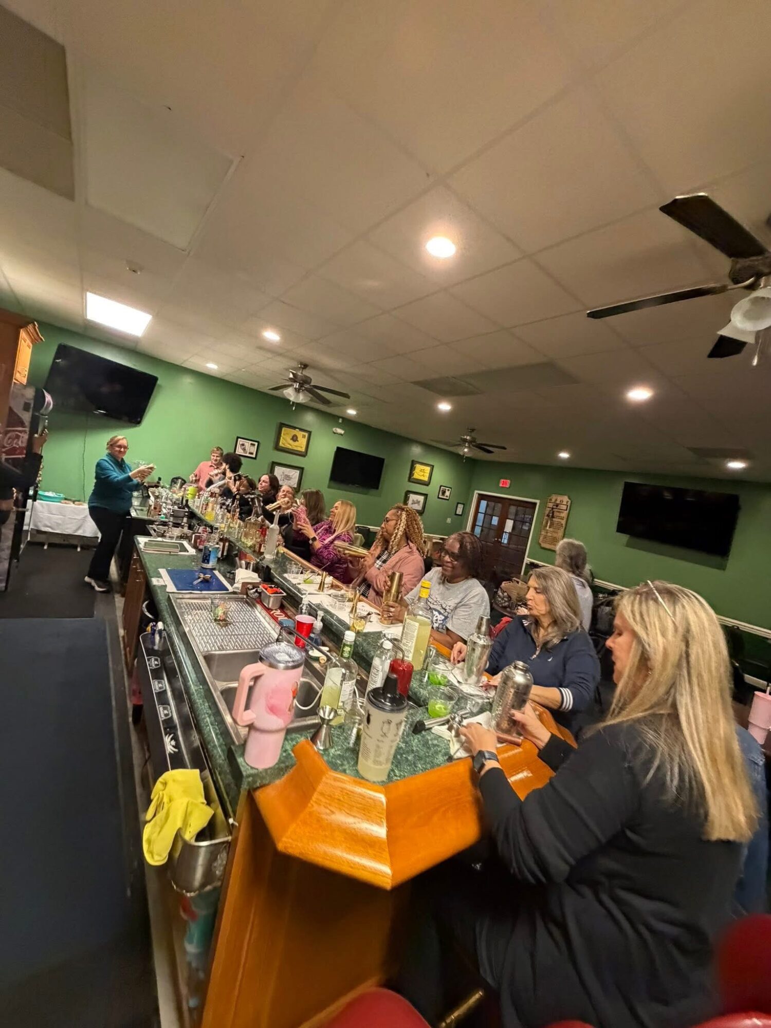 People gathered around a bar counter in a room with green walls, ceiling fans, and multiple television screens, socializing and drinking.