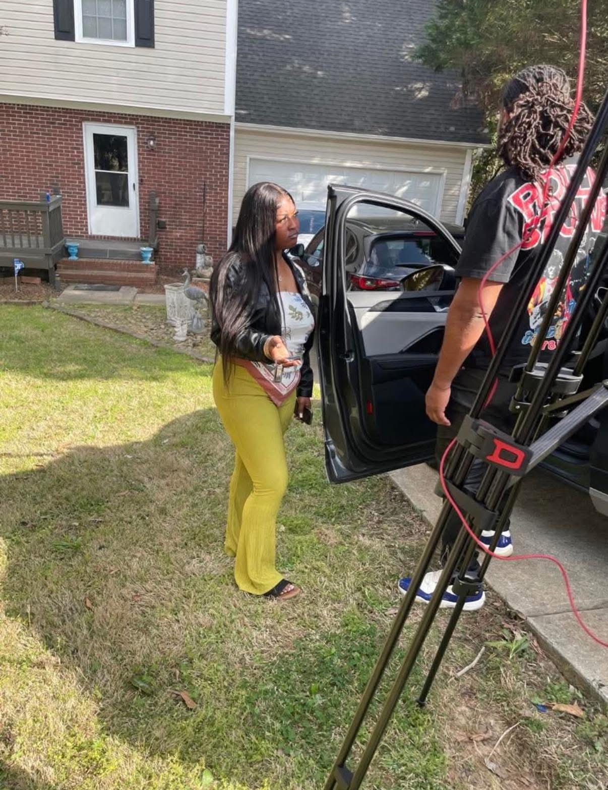 Two women standing outside near a car, one opening the car door, in a residential area with grass and houses