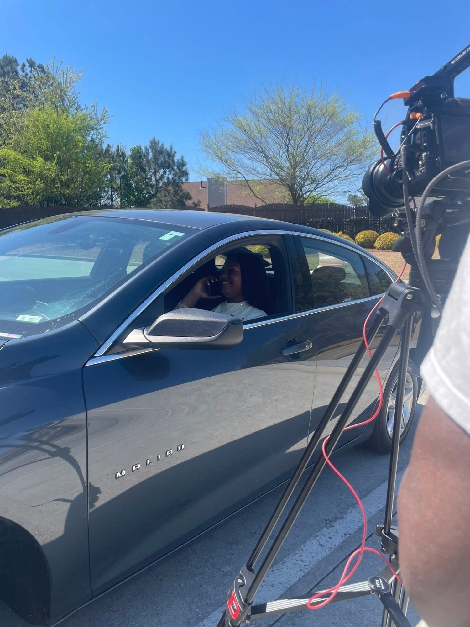 Person filming a dark gray Chevrolet car outdoors on a sunny day with trees and blue sky in background.