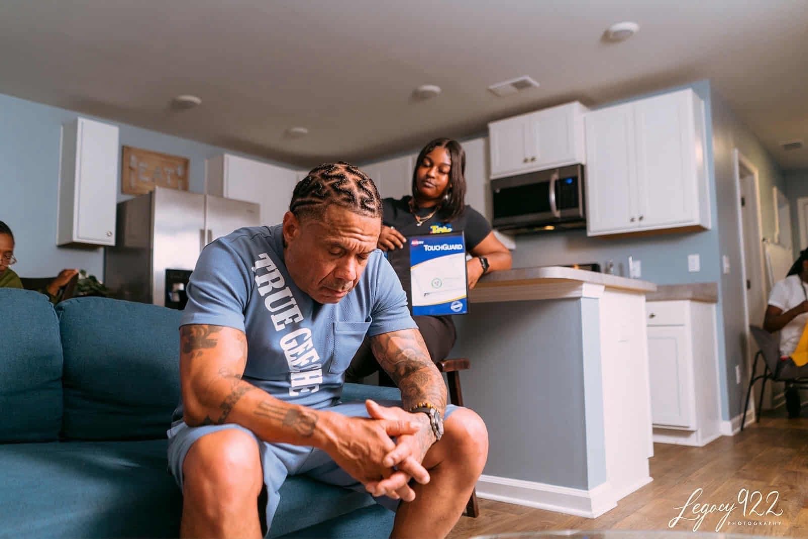 Man sitting on a couch in a kitchen, looking at his phone, with a woman standing behind the counter holding a box.