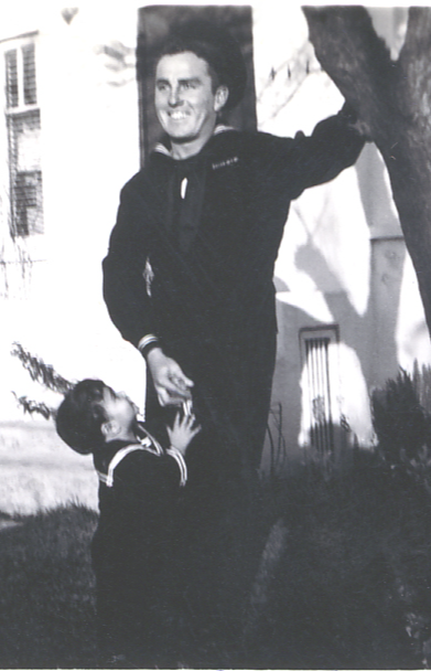 A man smiling outdoors with a child reaching up to him, trees and a building in background.