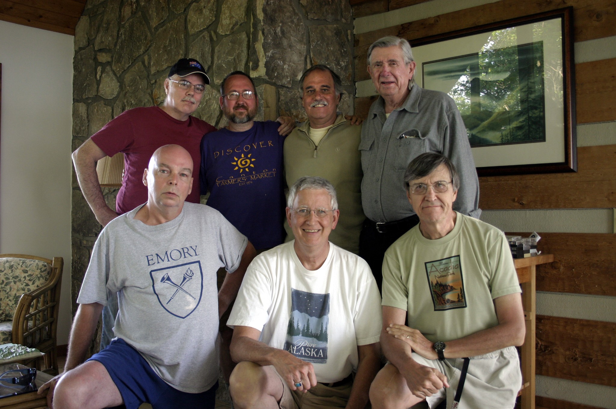 Seven people gathered indoors, some standing and some sitting, in front of a stone wall and framed picture.