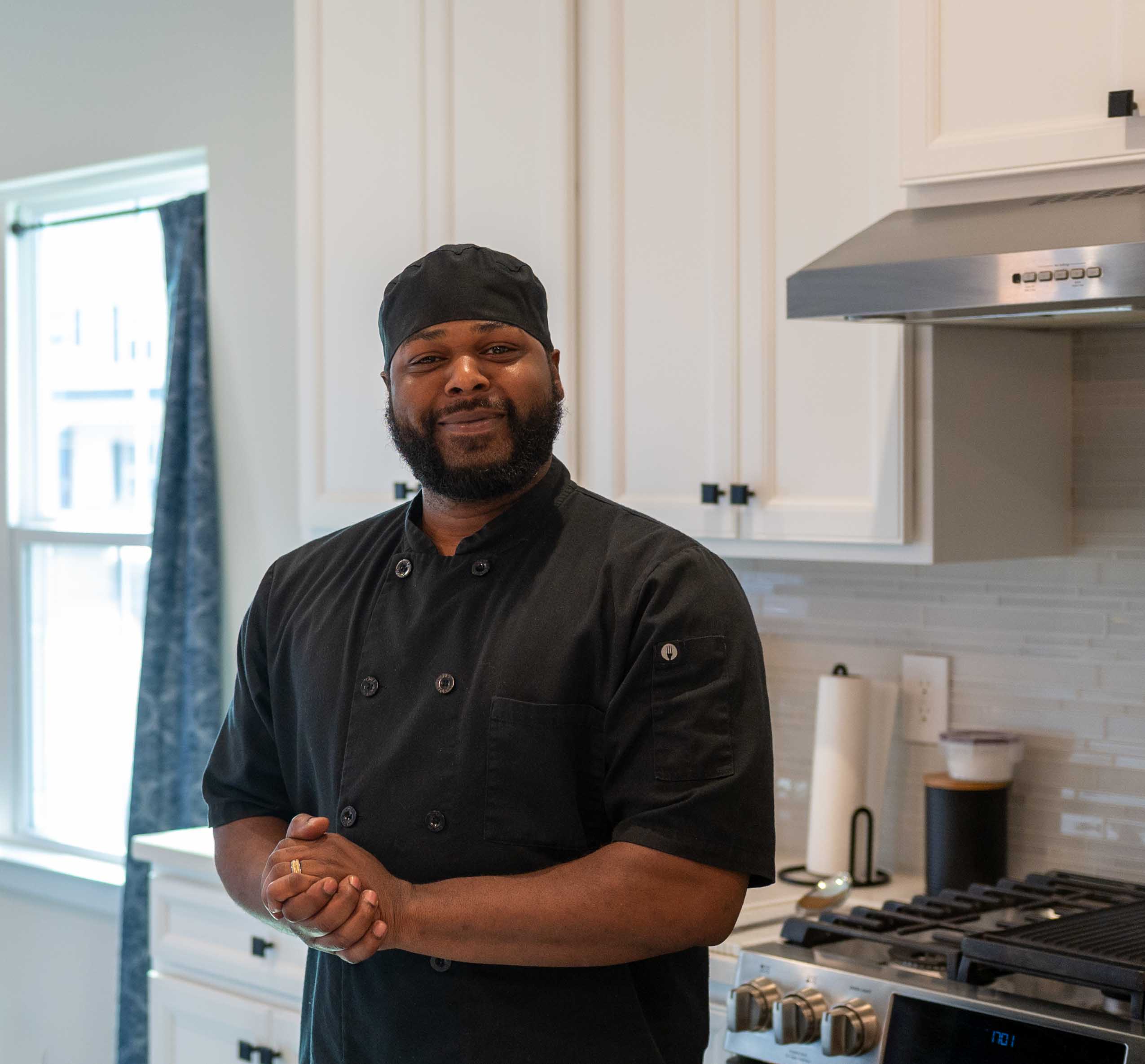 Chef in black uniform standing in a kitchen with white cabinets and a window, smiling with hands clasped.