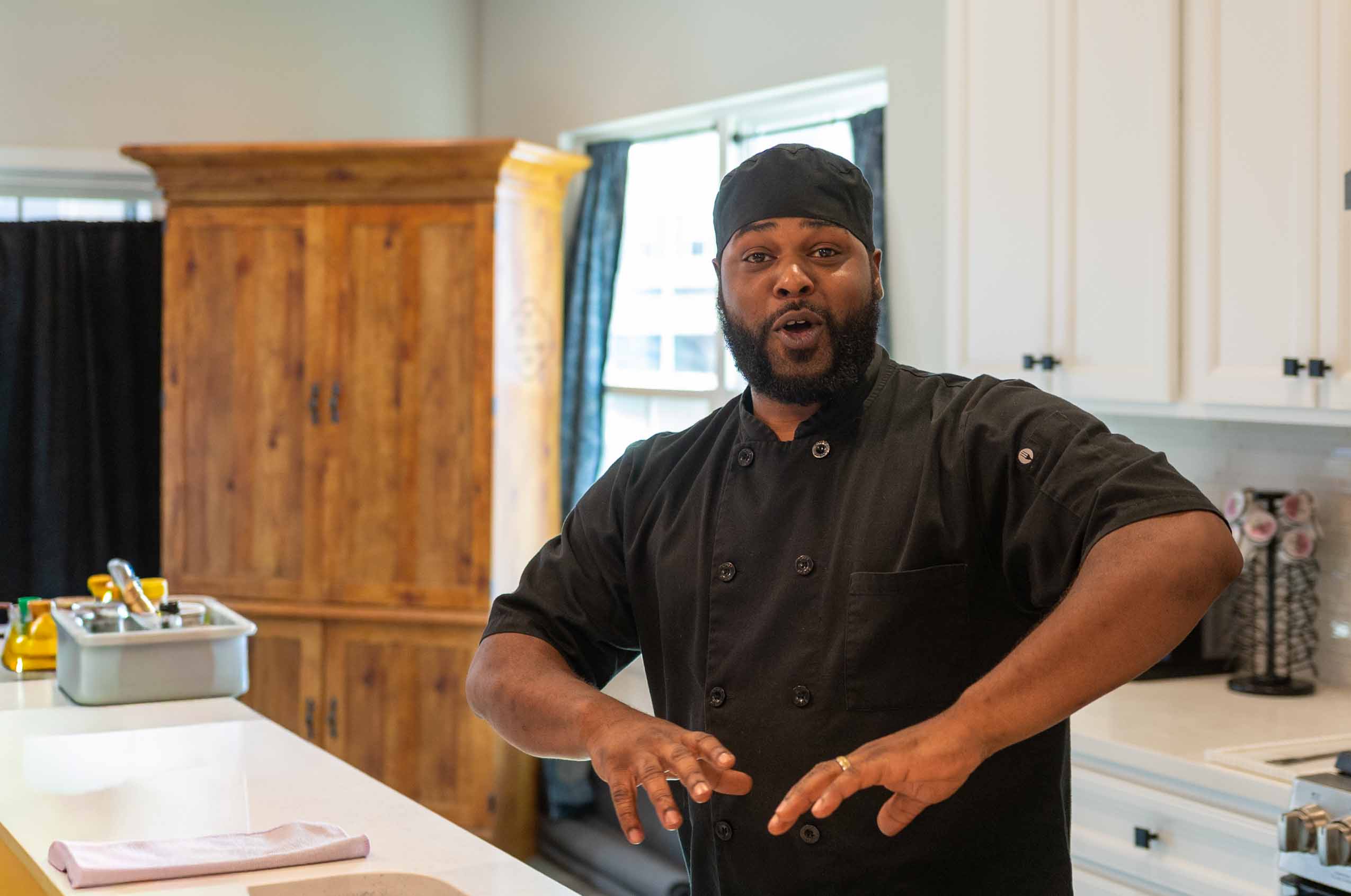 Chef in black uniform and head covering in a kitchen with white cabinets and wooden accents, gesturing with hands.