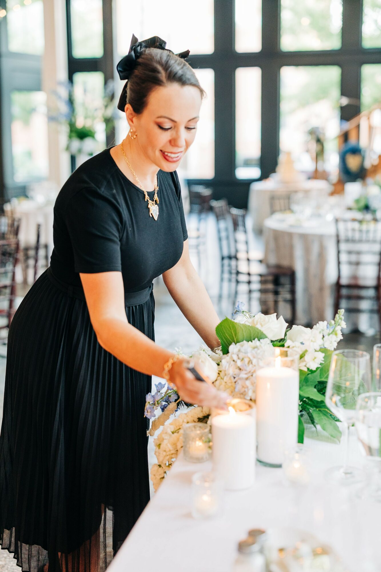 Woman arranging flowers on a table with candles in a bright, elegant room with large windows.