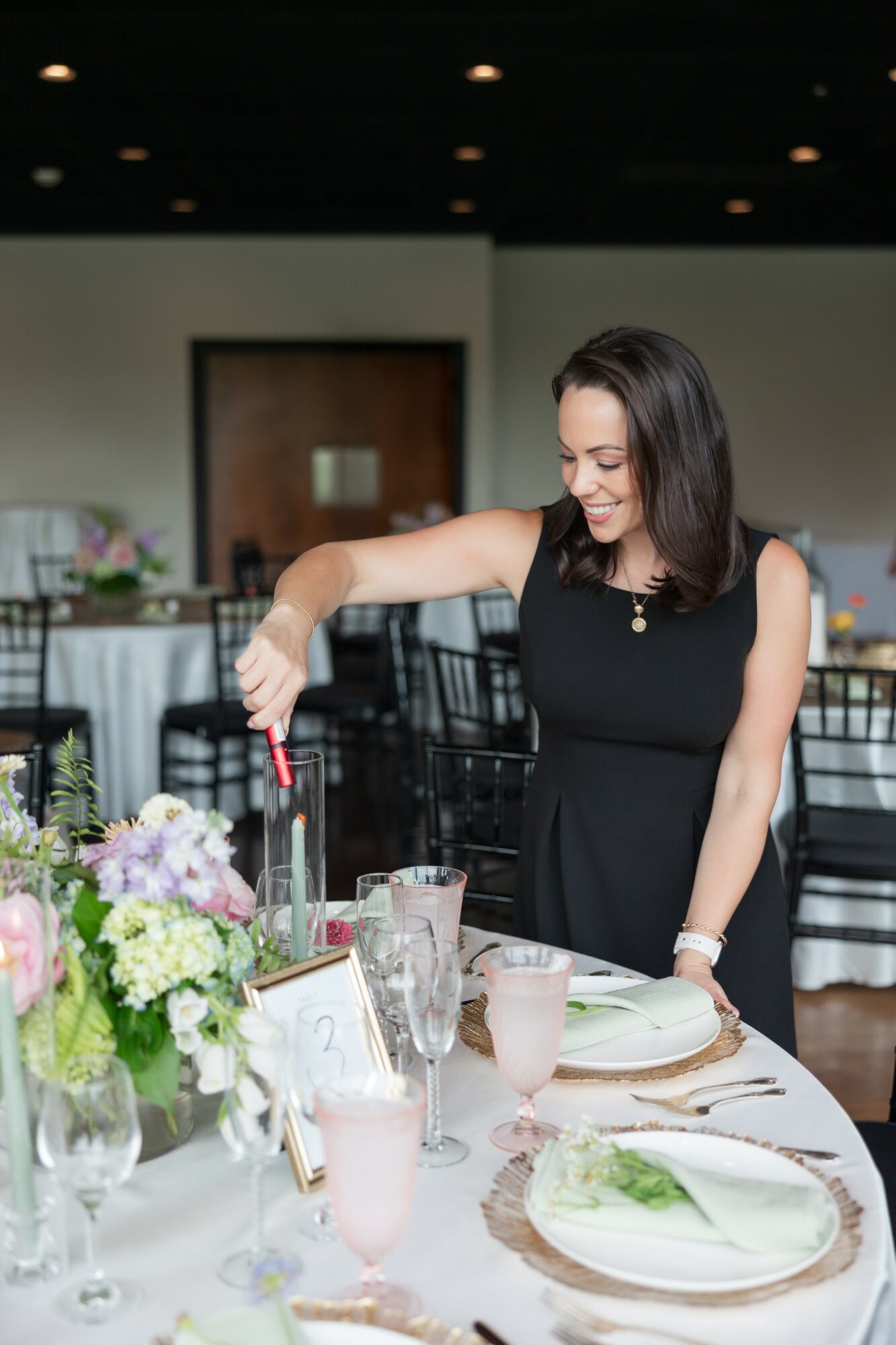 Woman in black dress setting a table with flowers and glassware, smiling, in a dining room.