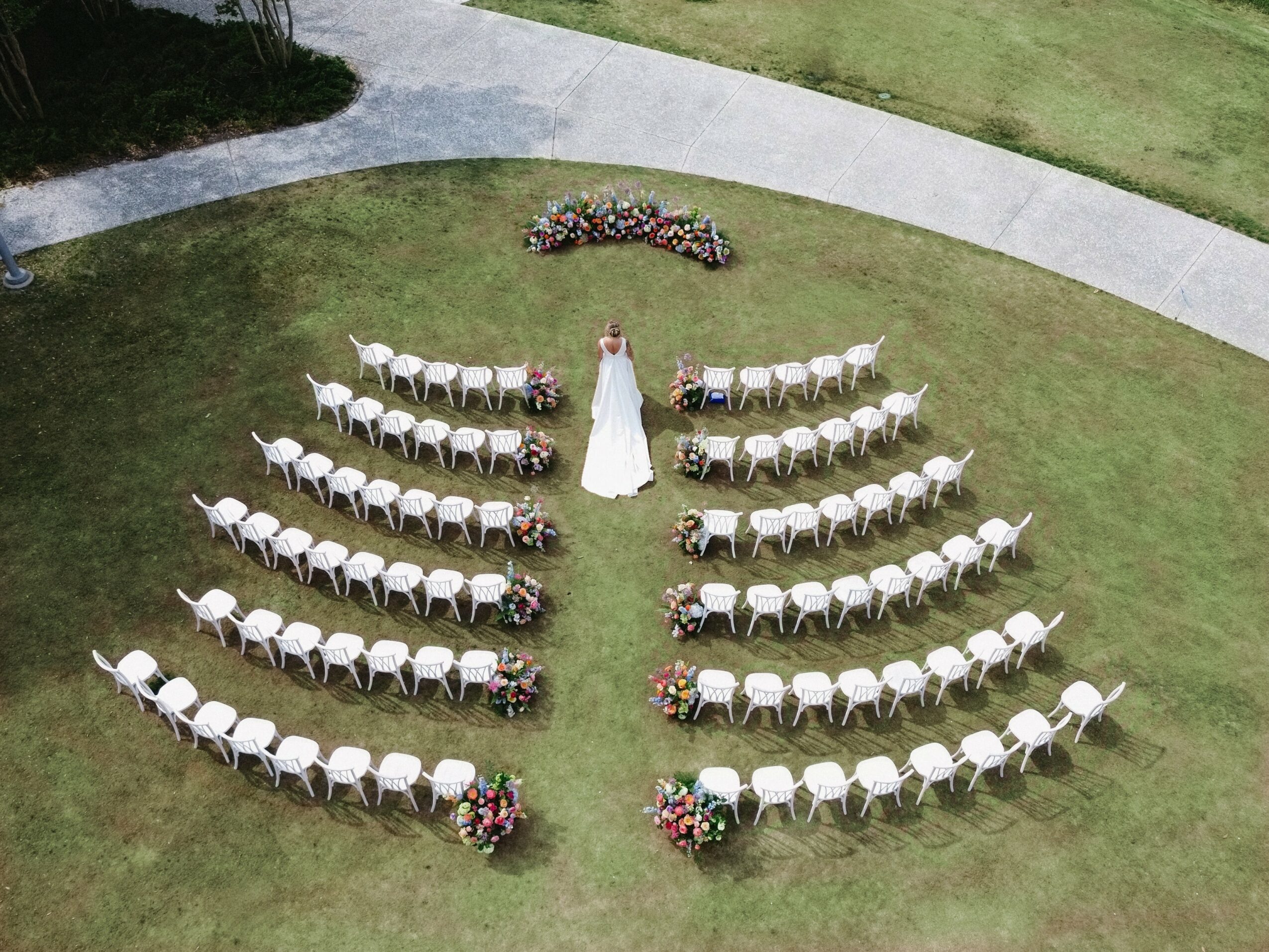 Wedding ceremony setup with chairs arranged in a semi-circle around a bride in a white dress, on a grassy area.