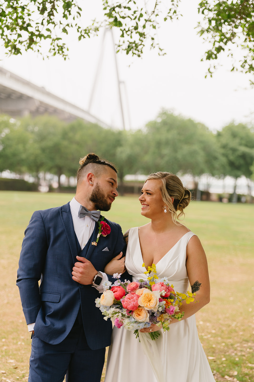 A couple in wedding attire outdoors, the man in a blue suit and the woman in a white dress holding a bouquet, smiling at each other.
