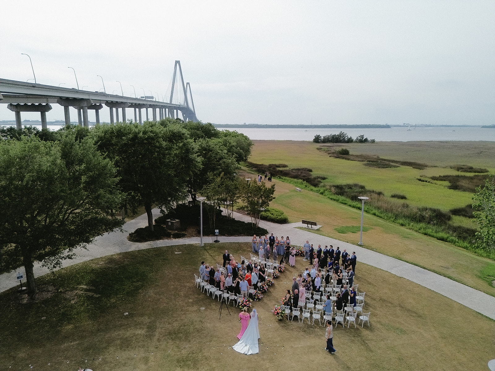 People gathered outdoors near trees and a pathway, with a bridge and water in the background, during daytime.
