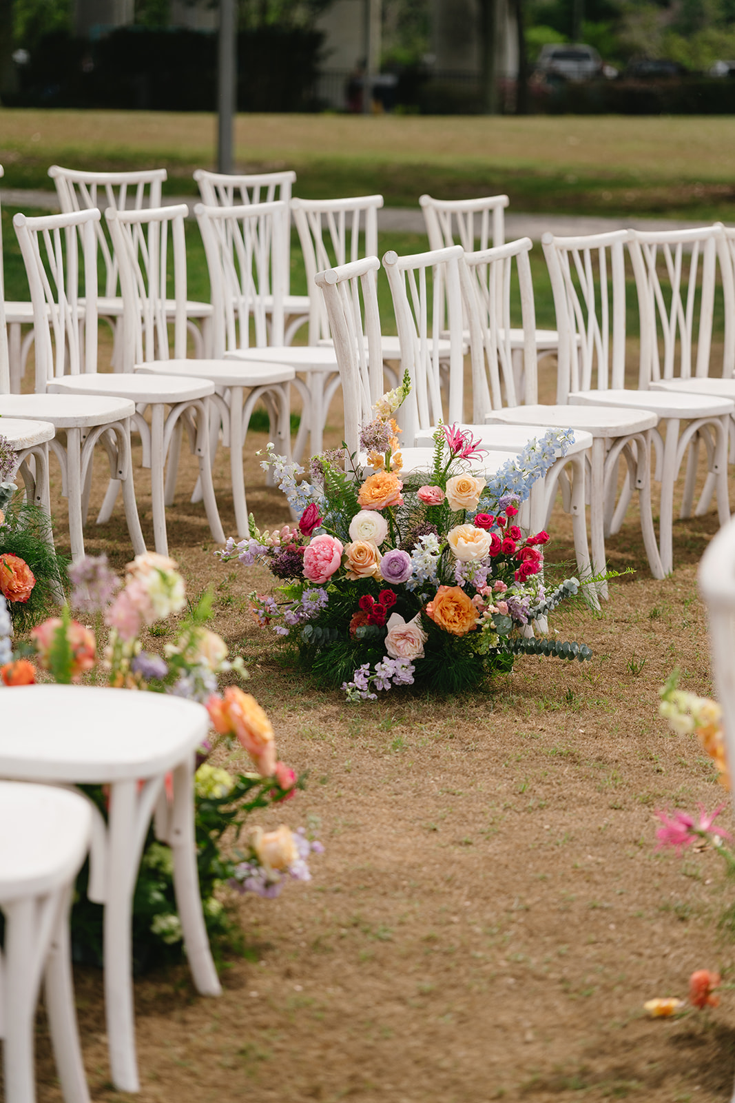White chairs arranged outdoors with floral arrangements on the ground, grass and trees in the background.