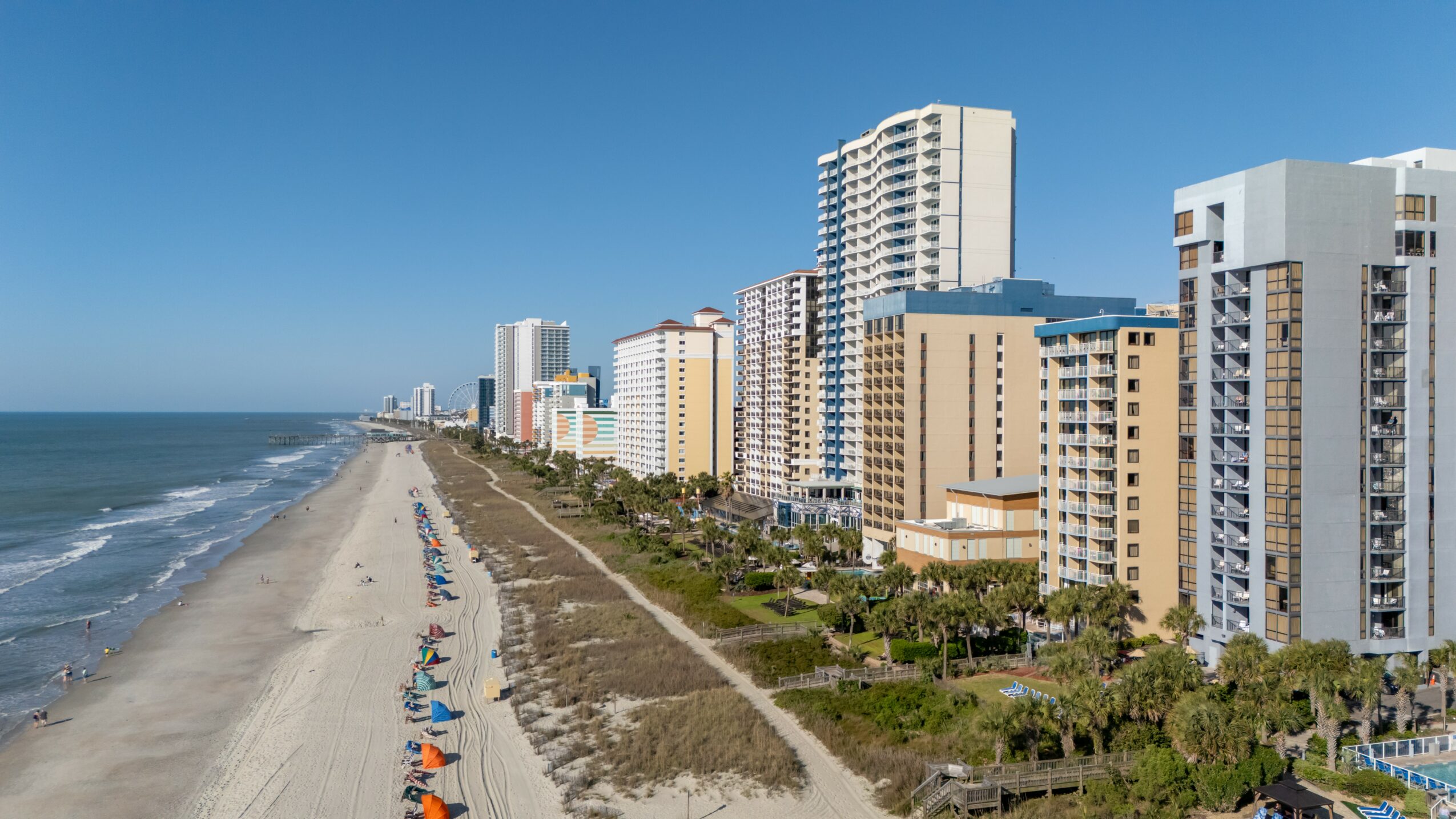 Beach with high-rise buildings along the shoreline, clear blue sky, and ocean waves, with some beach chairs and umbrellas visible.