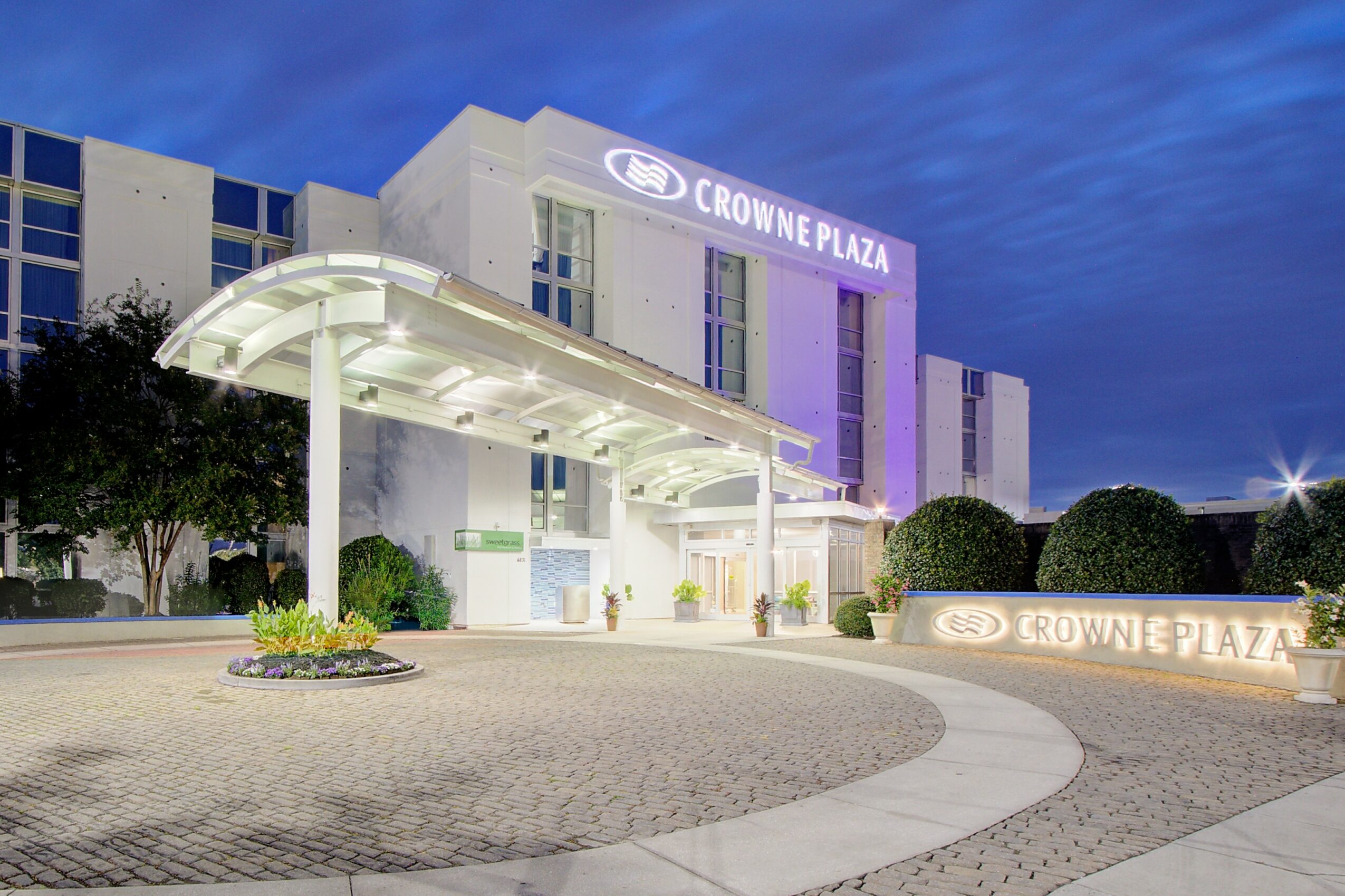 Crowne Plaza hotel exterior at dusk with illuminated signage and entrance canopy, surrounded by trees and a circular driveway.
