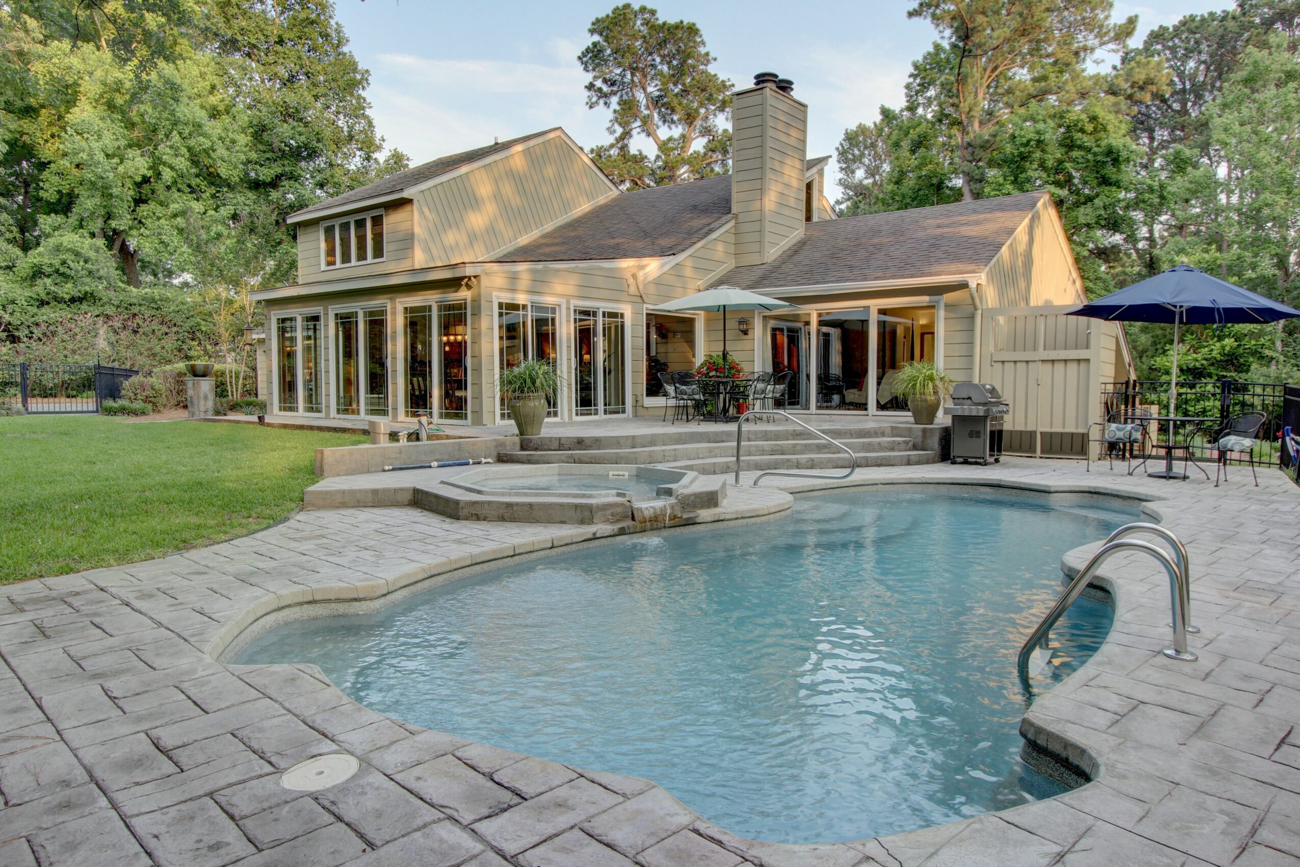 Backyard with a swimming pool, patio, house with large windows, and outdoor furniture, surrounded by trees and greenery.