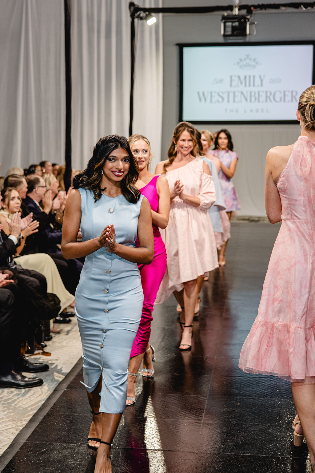 Women walking on runway during fashion show, audience seated on sides, screen displaying designer's name in background.
