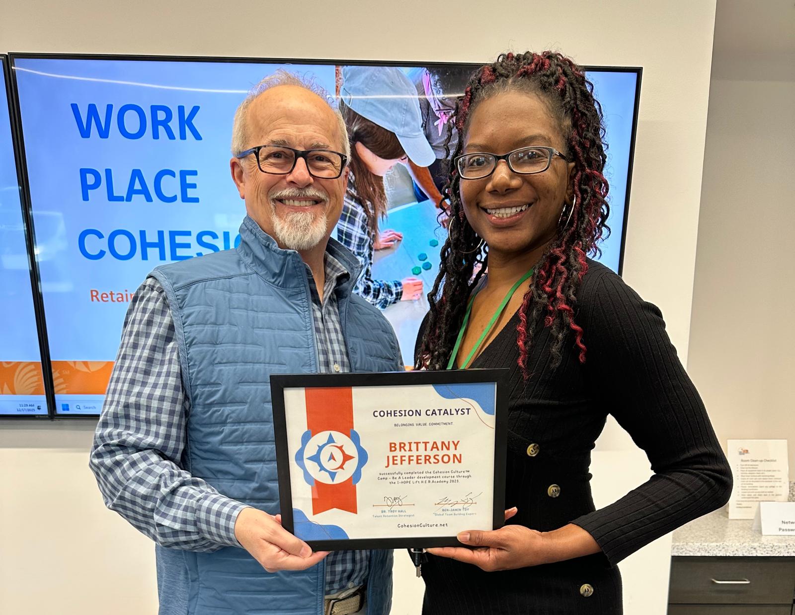 Two people smiling, one holding a certificate, in front of a screen with text about workplace cohesion.
