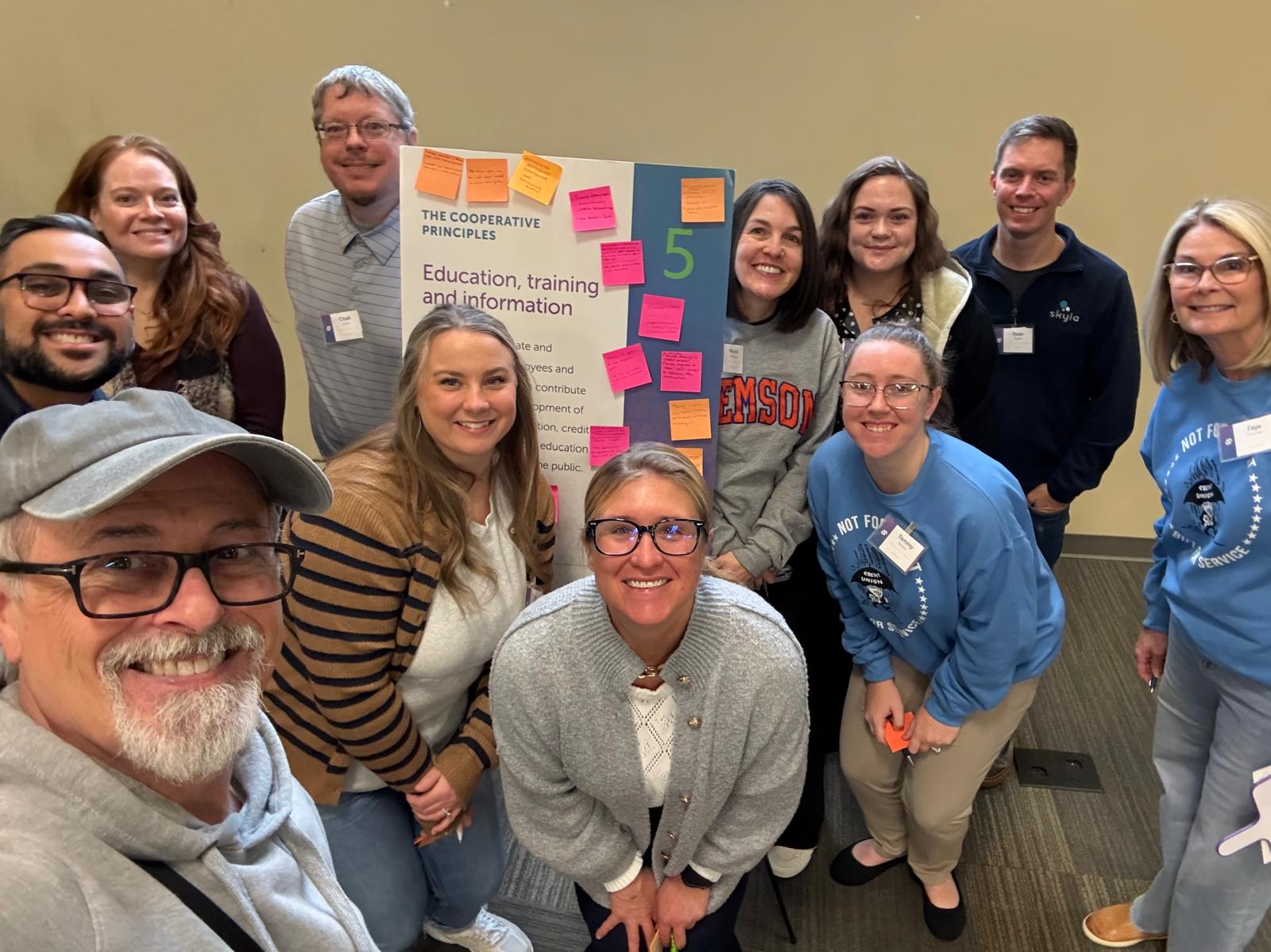 Group of ten people smiling, standing around a colorful poster with sticky notes, in a room with beige walls.