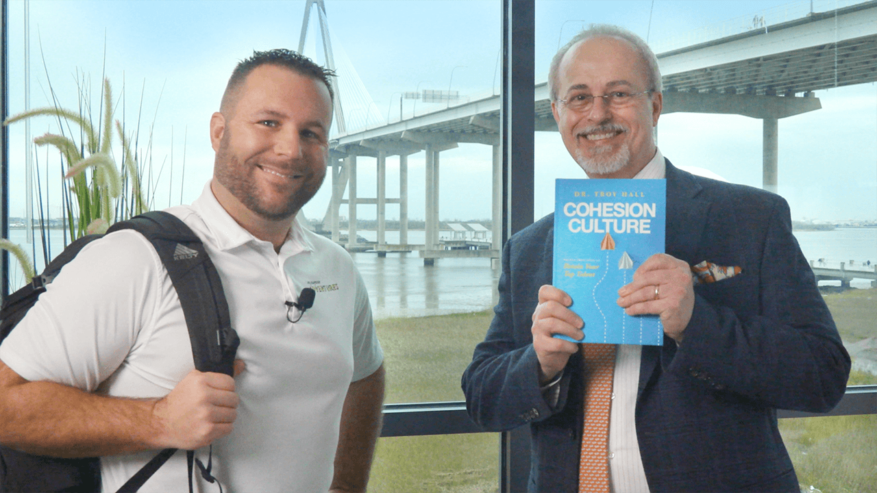 Two men standing indoors near a window, one holding a book titled 'Cohesion Culture', smiling.