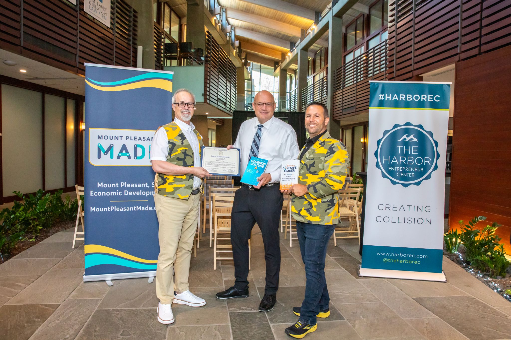 Three men standing indoors, holding certificates, with banners in the background, smiling for the photo.