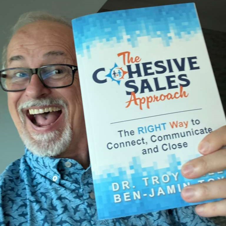 Smiling man with glasses holding a book titled 'The Cohesive Sales Approach' in front of his face.