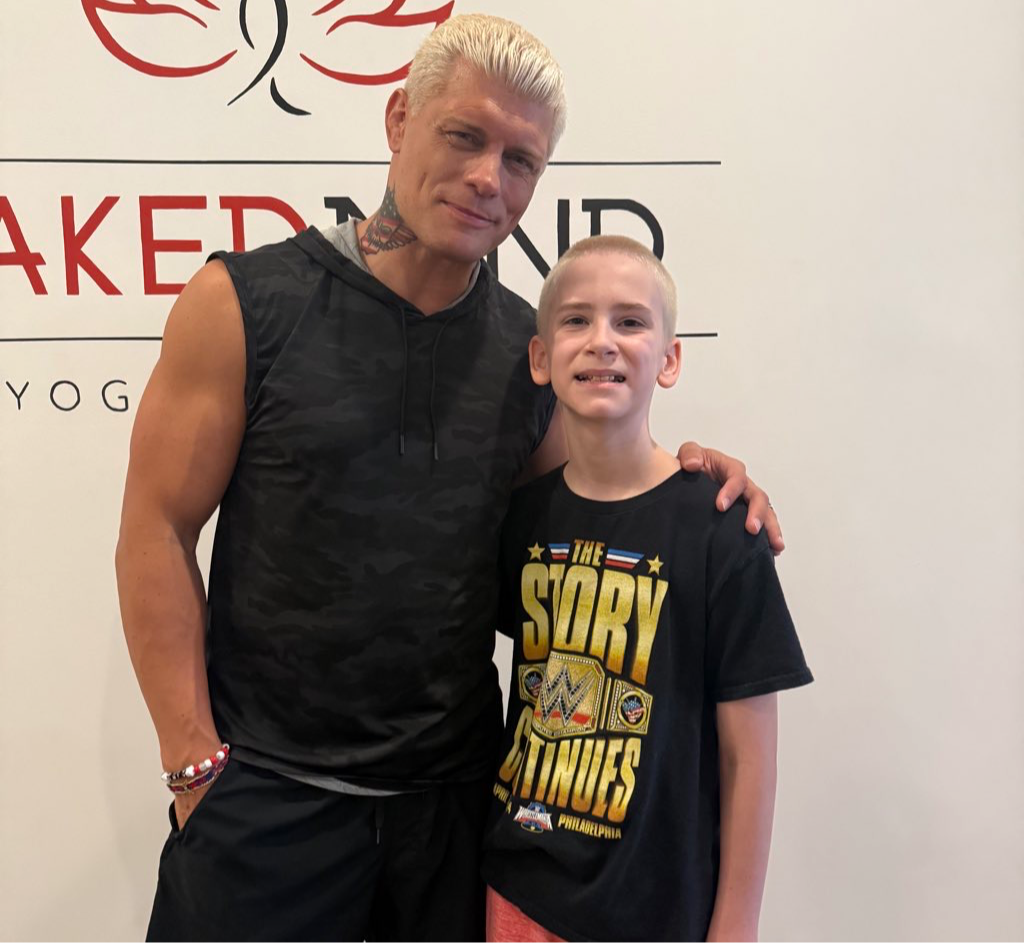 Man with platinum blonde hair and a young girl with a shaved head stand together indoors, smiling. The man has his arm around the girl.