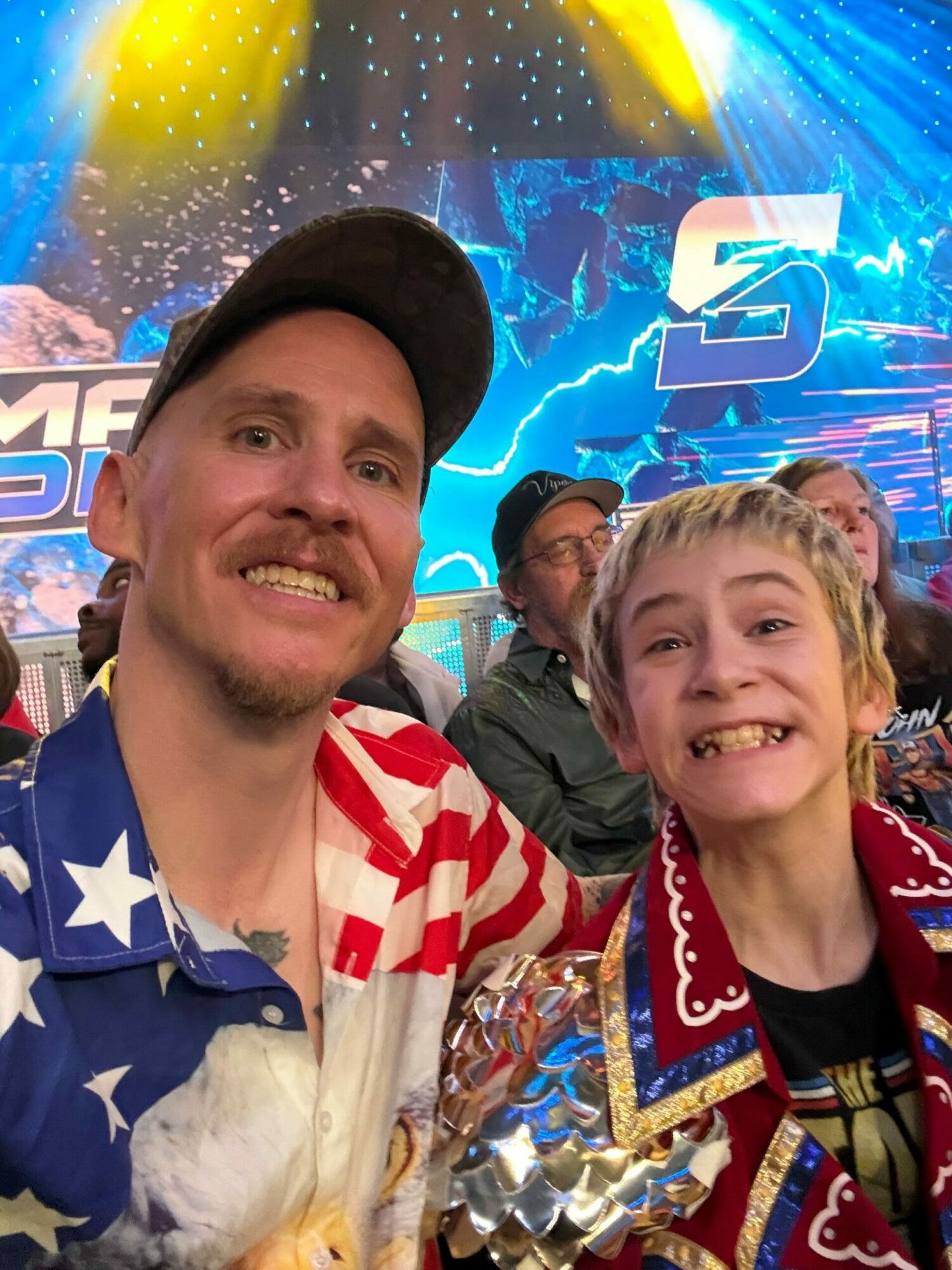 Two smiling people at an event with a colorful, illuminated background, one wearing a blue star-patterned shirt and the other in a red and white jacket.