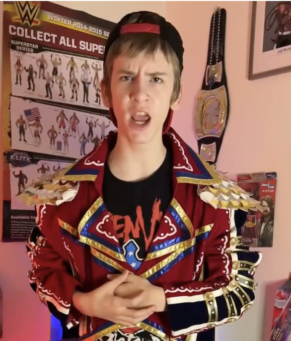 Young person with short hair wearing a red and gold jacket, standing indoors with posters and a championship belt behind them.