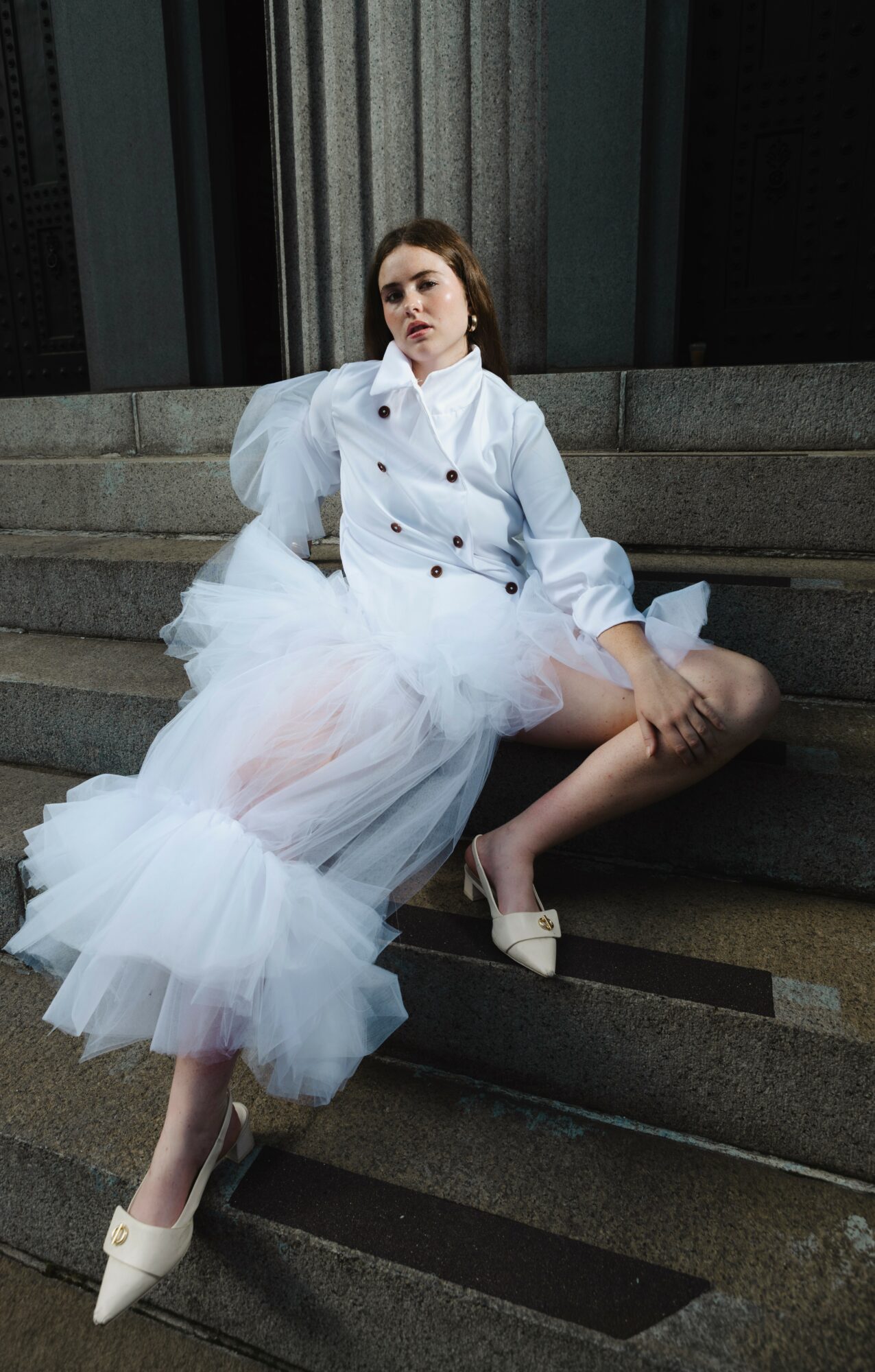 Woman in a white jacket and tulle skirt sitting on a stone ledge, with one leg extended and wearing white heels.