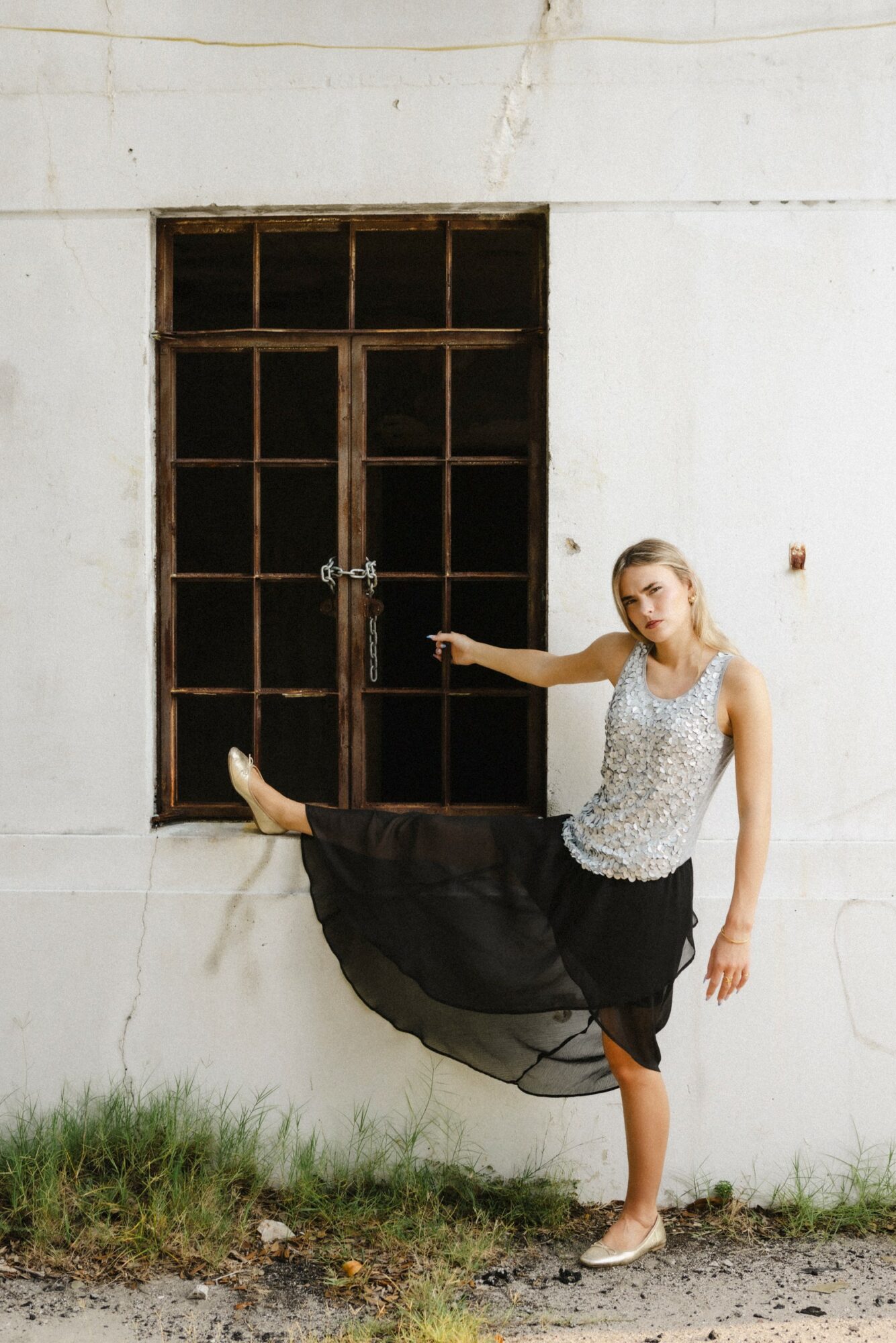 Woman in a dress and skirt poses with one leg raised against a white wall and window, outdoors.