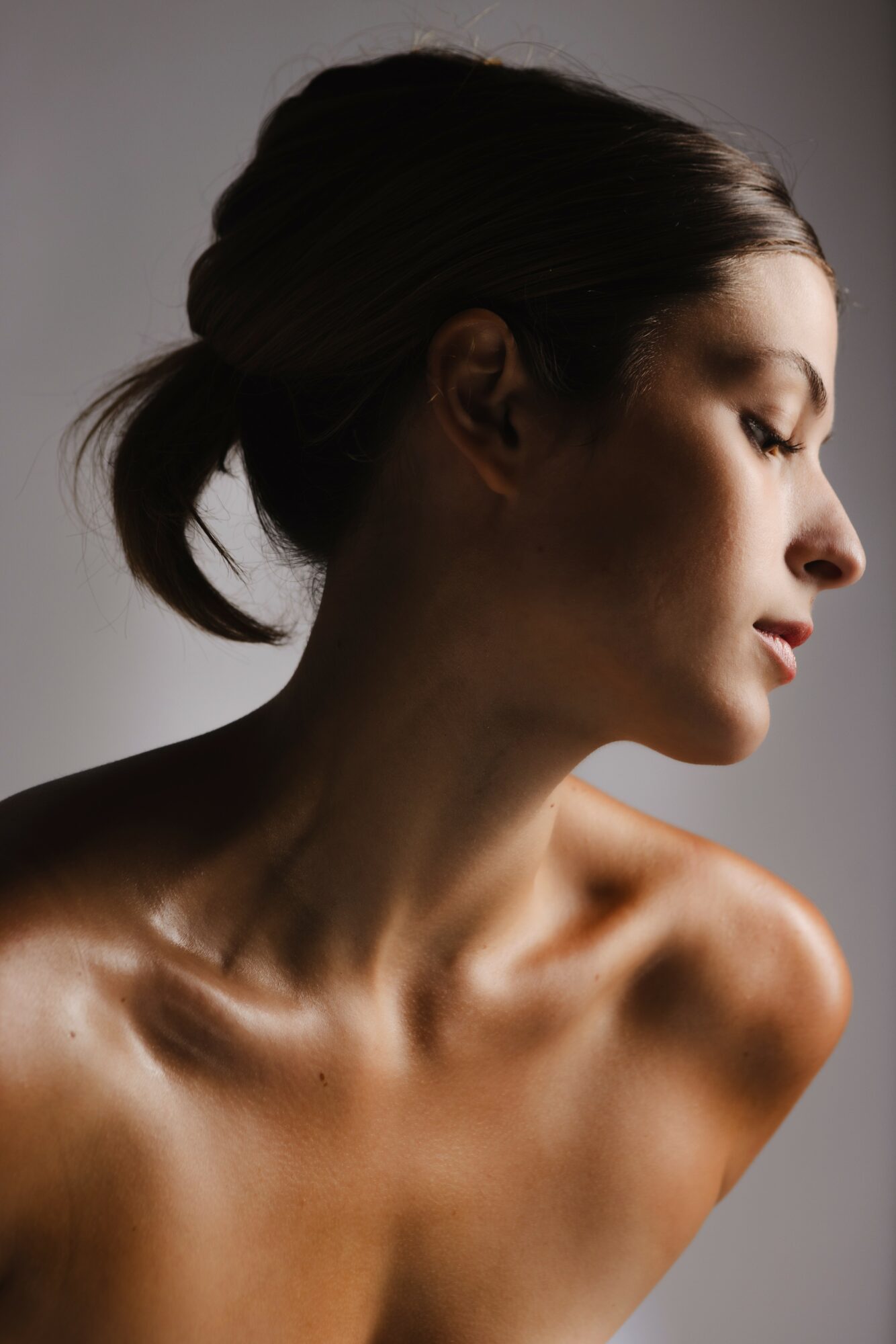 Profile of a woman with her hair in a bun, looking downward, with bare shoulders and collarbone visible.