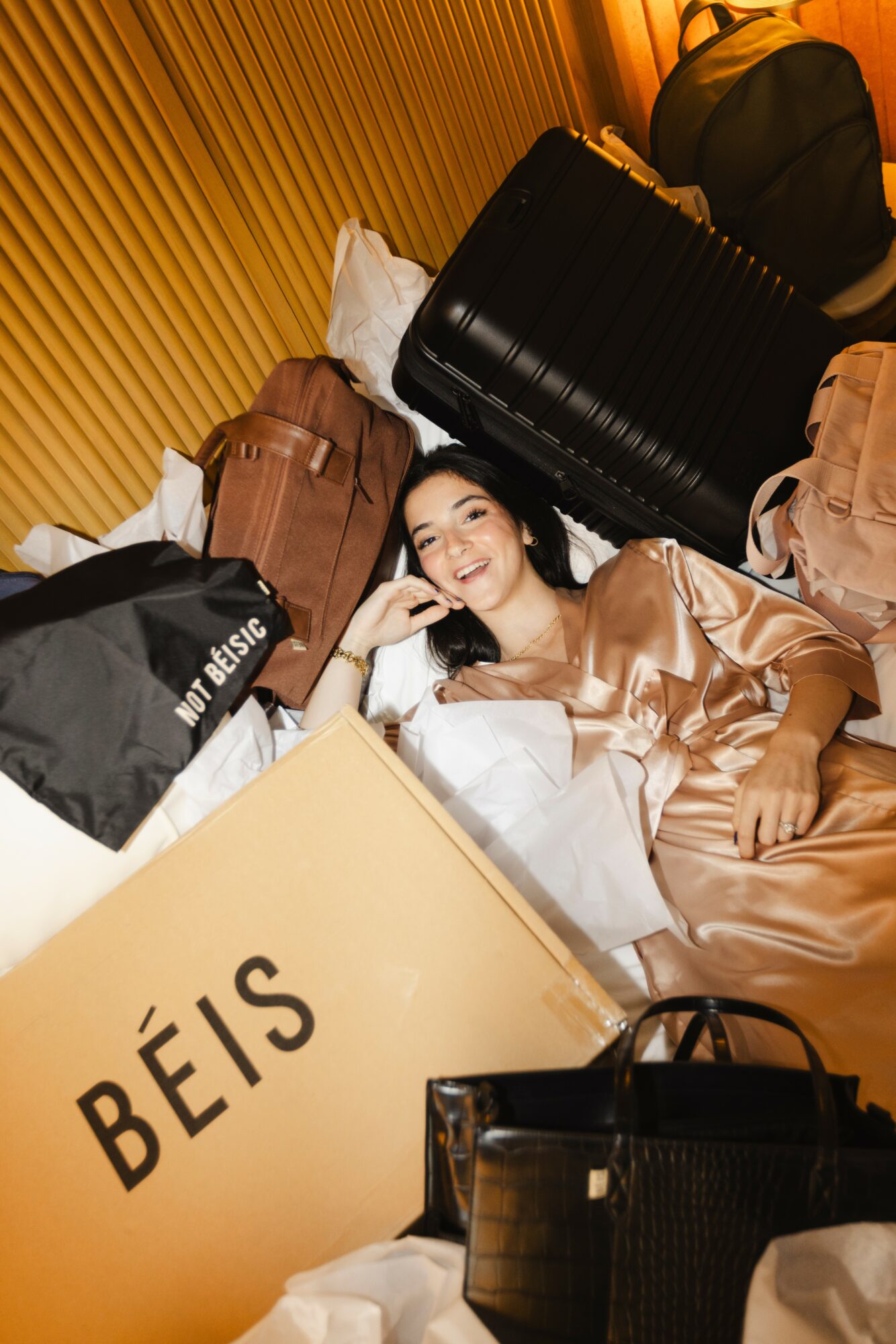 Woman lying on bed surrounded by luggage and shopping bags, smiling and resting her head on her hand.