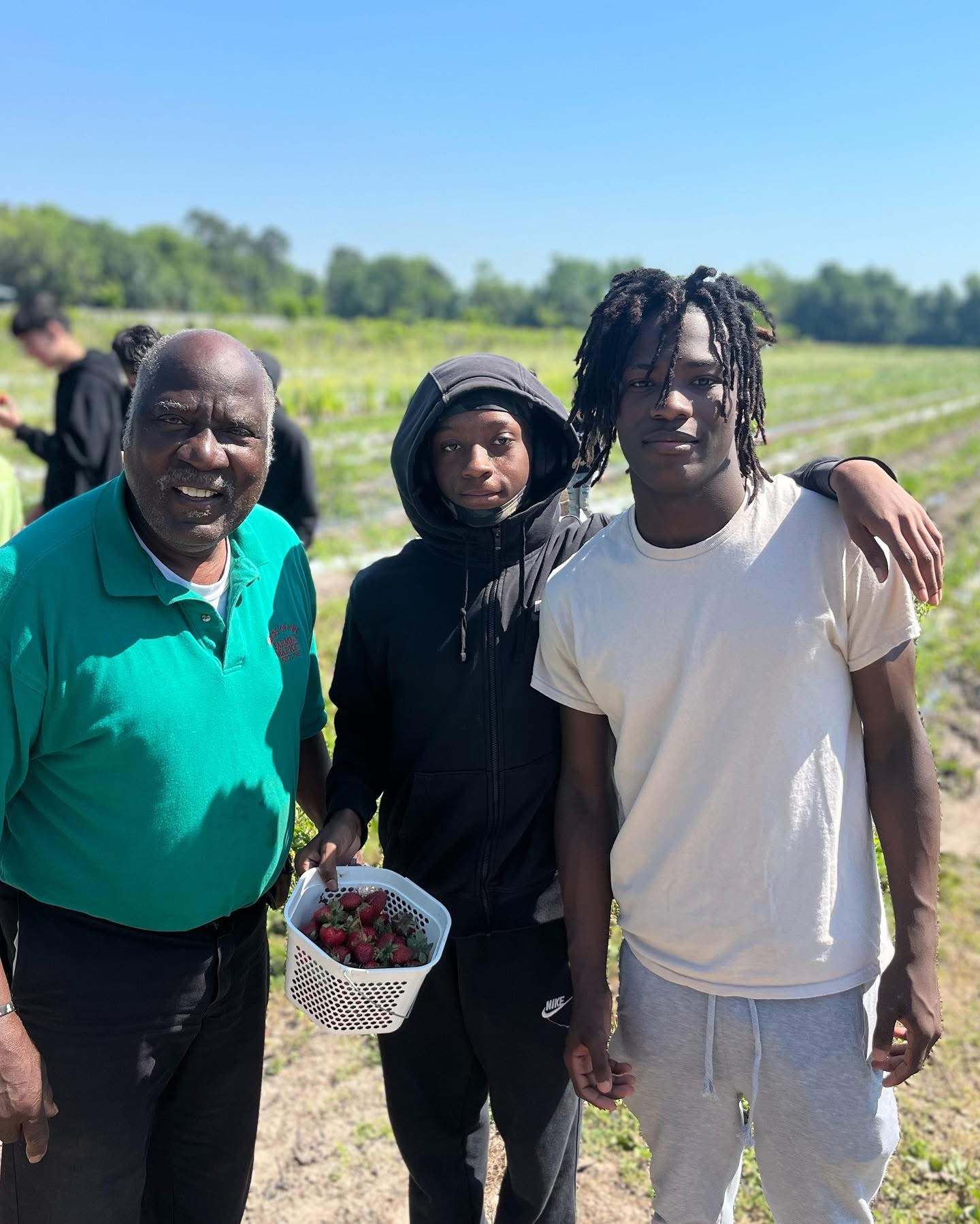 Three people standing outdoors in a field, one holding a basket of strawberries, with trees and blue sky in background.